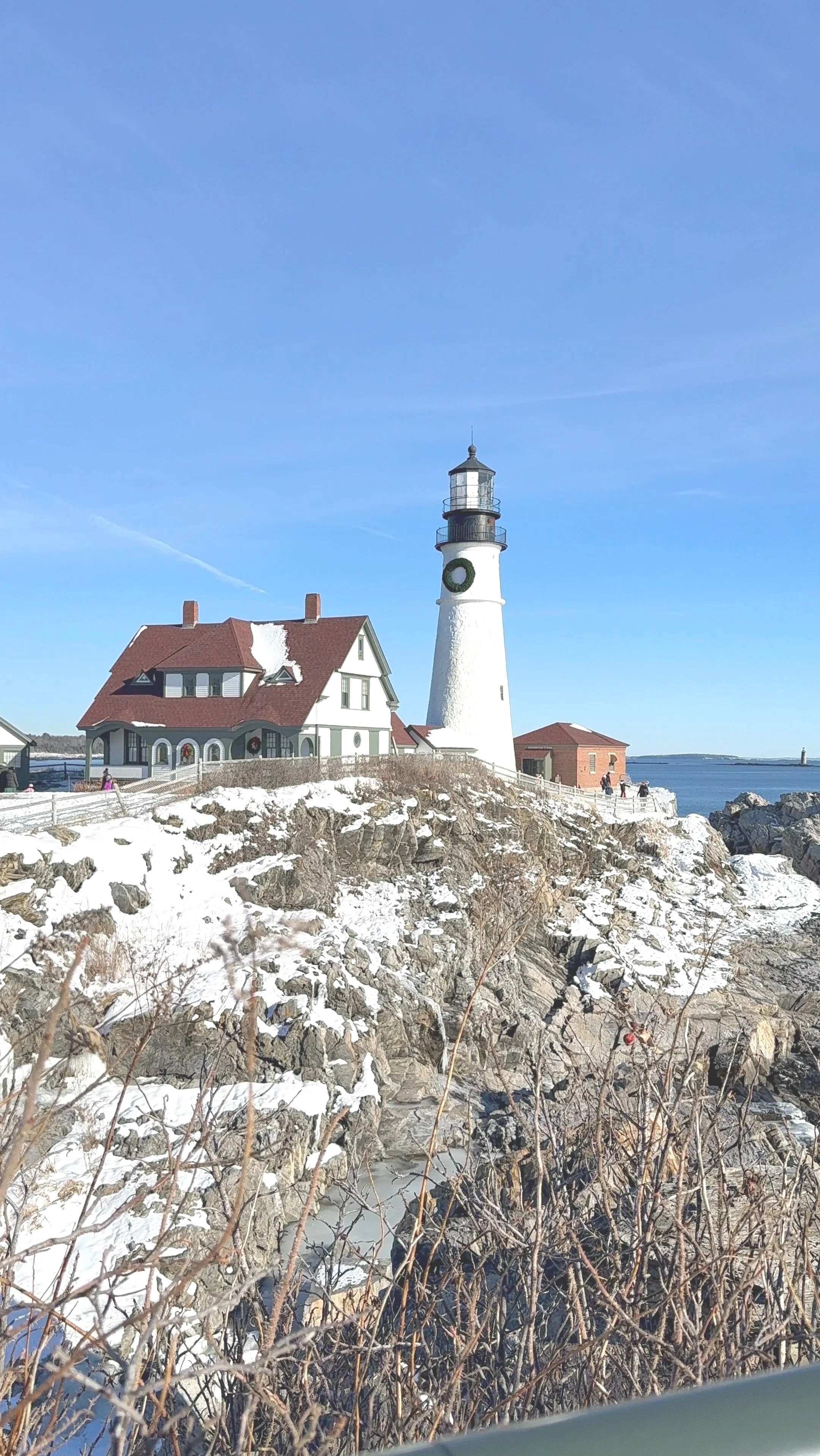 Portland Head Light lighthouse and keeper's quarters surrounded by snow on the rocky Maine coastline under a clear blue sky.