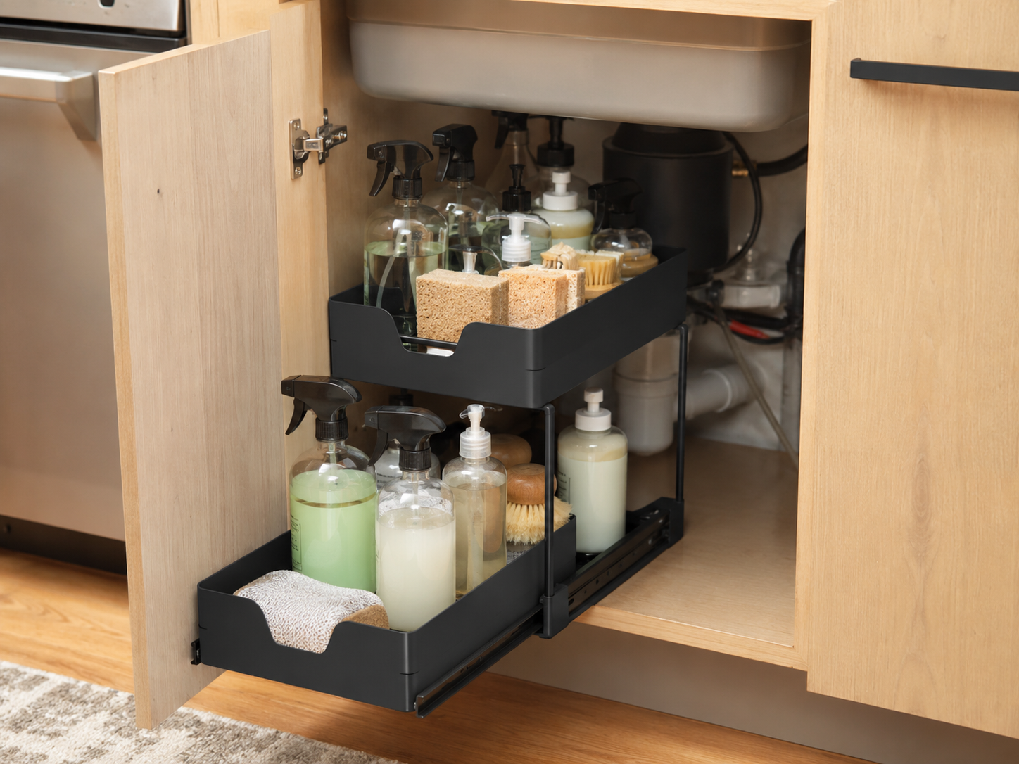 Two-tier sliding under-sink organizer holding cleaning sprays, soap bottles, scrub brushes, sponges, and cloths inside a wood kitchen cabinet.