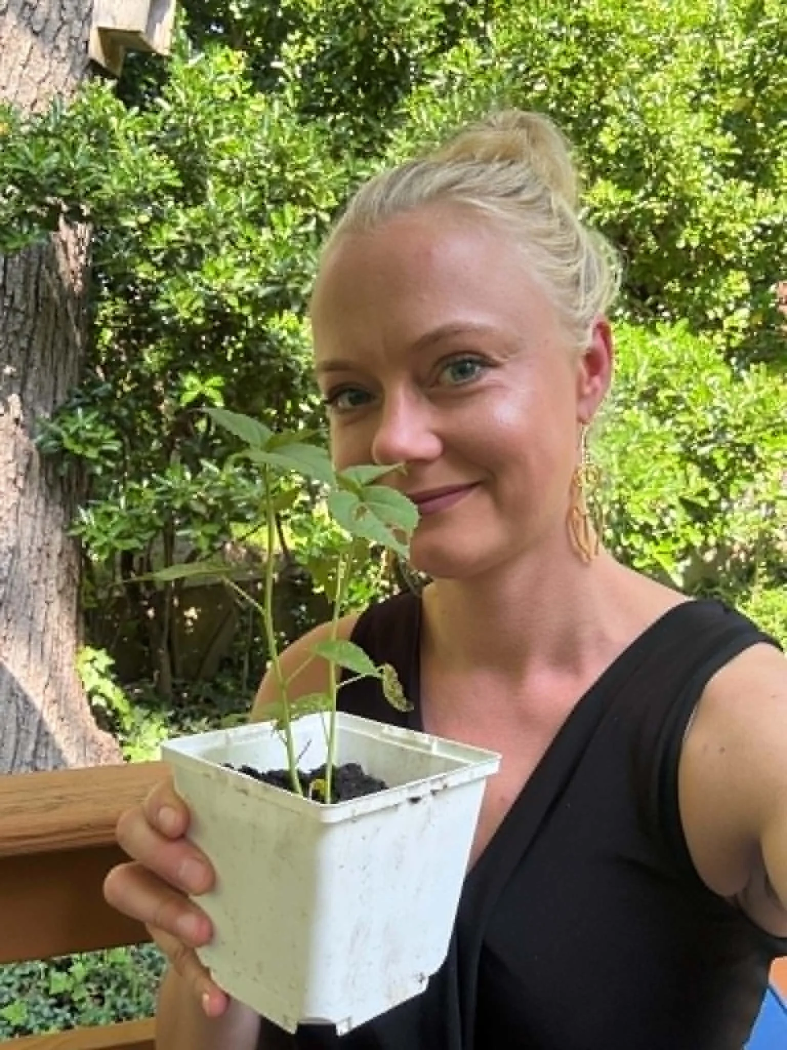 A woman with blonde hair in a bun, wearing earrings and a black top, smiling while holding a white container with small green plants inside, outdoors with green trees in the background.