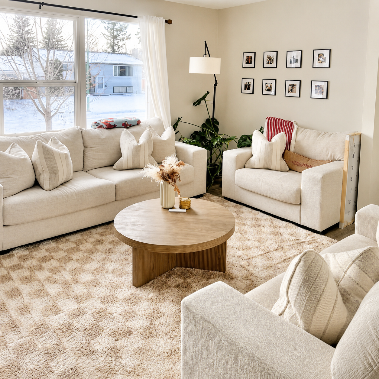 A cozy living room with beige sofas, decorative pillows, a wooden coffee table with a vase and candles, a large window with white curtains, a potted plant, wall art, and a plush rug.