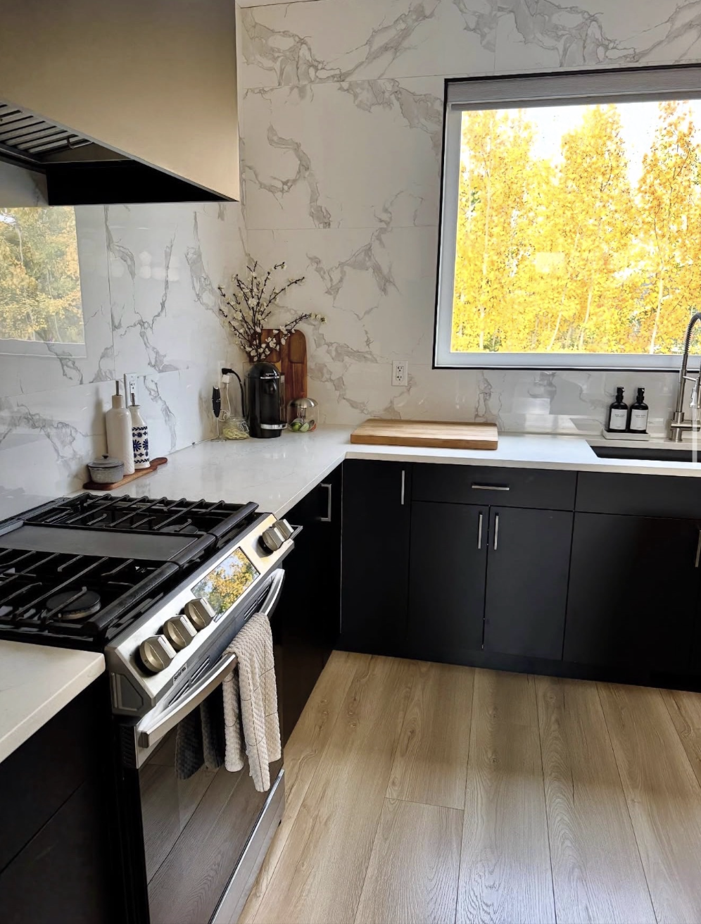 Modern kitchen with white marble backsplash, black cabinets, and a window showing yellow trees outside. Has a stove, coffee station, and kitchen accessories.