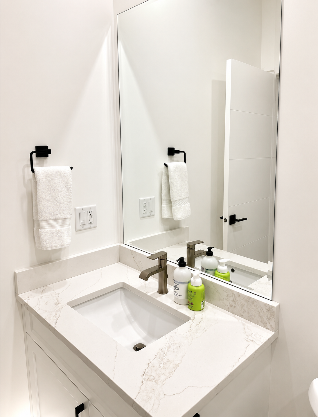 Bathroom vanity with a white marble countertop, undermount sink, and a large mirror. Hand towels on black towel rings, soap and lotion bottles on the counter, and a door reflected in the mirror.