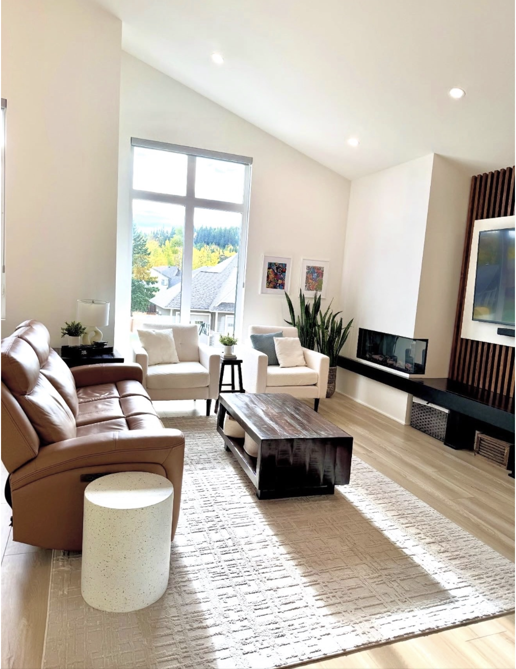 Modern living room with beige leather couch, white armchairs, wooden coffee table, large window, plants, television, and artwork on the wall.