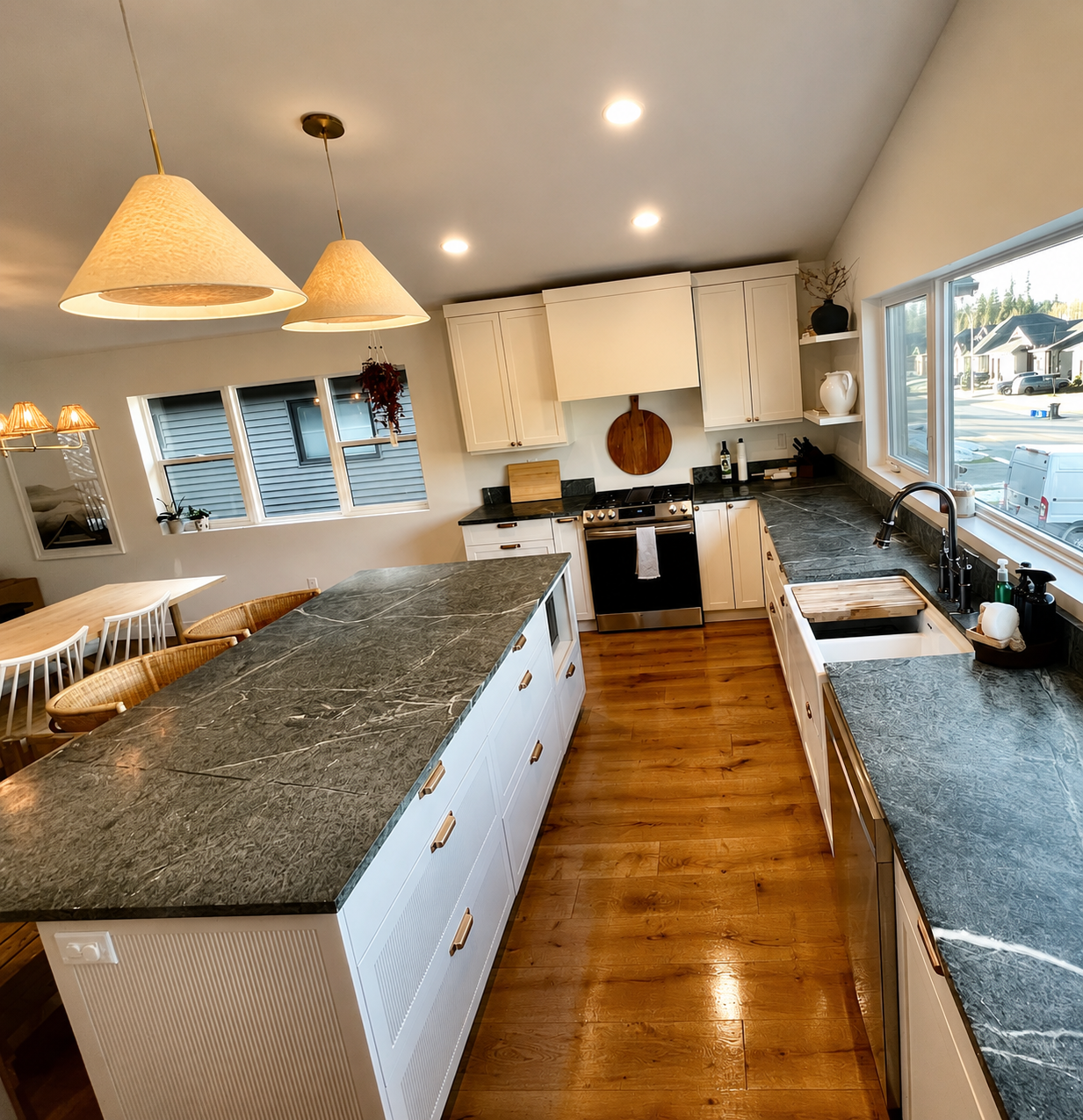Modern kitchen with large island, black countertops, white cabinets, stainless steel stove, and hardwood floors, with a dining area and large windows letting in natural light.