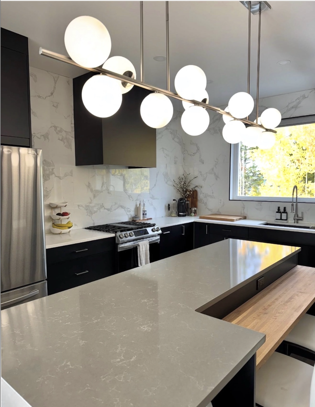 Modern kitchen with black cabinets, white marble backsplash, stainless steel refrigerator, and a large countertop with lighting fixture above, a window with trees outside, and kitchen essentials on the counters.