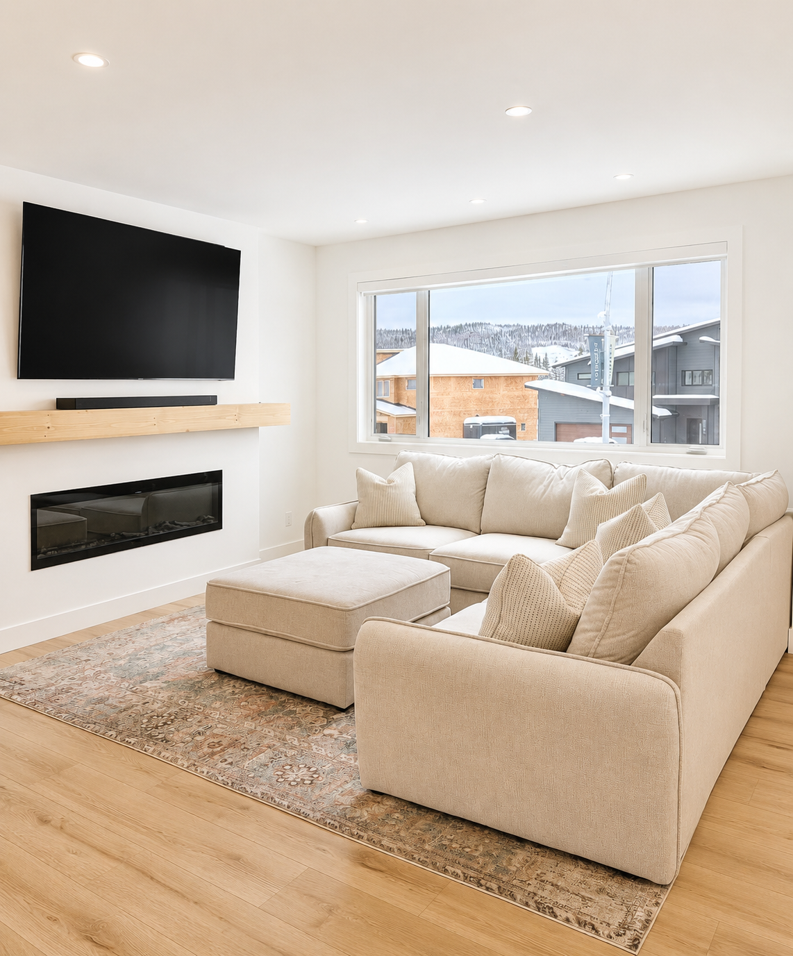 Living room with beige sectional sofa, matching ottoman, patterned area rug, large window showing snow-covered landscape, wall-mounted TV, electric fireplace, and soundbar.