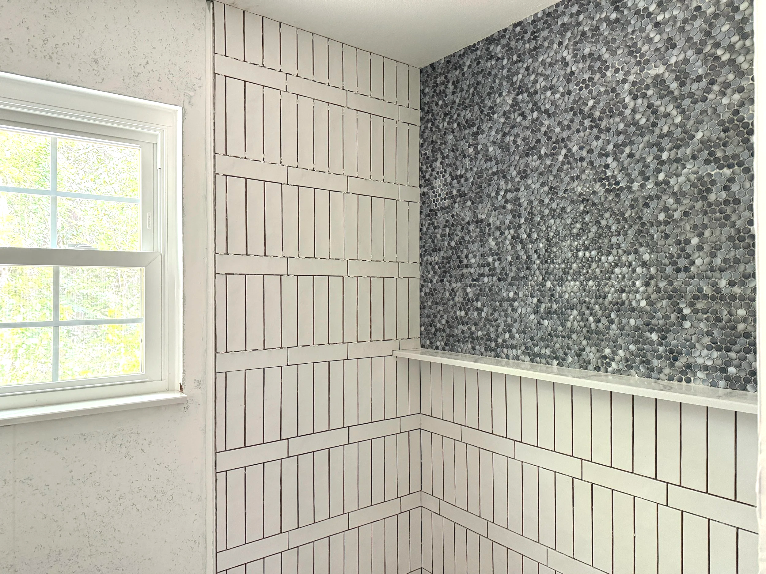 A section of a bathroom wall with white rectangular tiles and dark pebble mosaic tiles beneath a window, with white trim and an outdoor view of green foliage.
