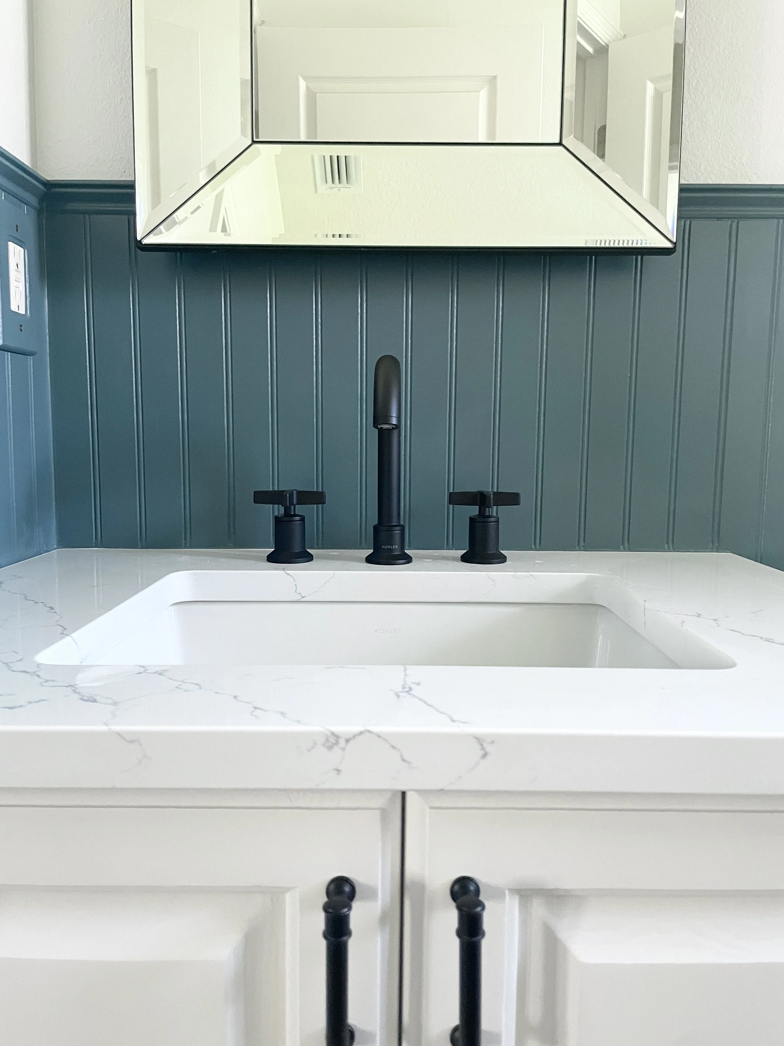A kitchen sink with white marble countertop, dark green beadboard wall, black faucet, and a large mirror above the sink.