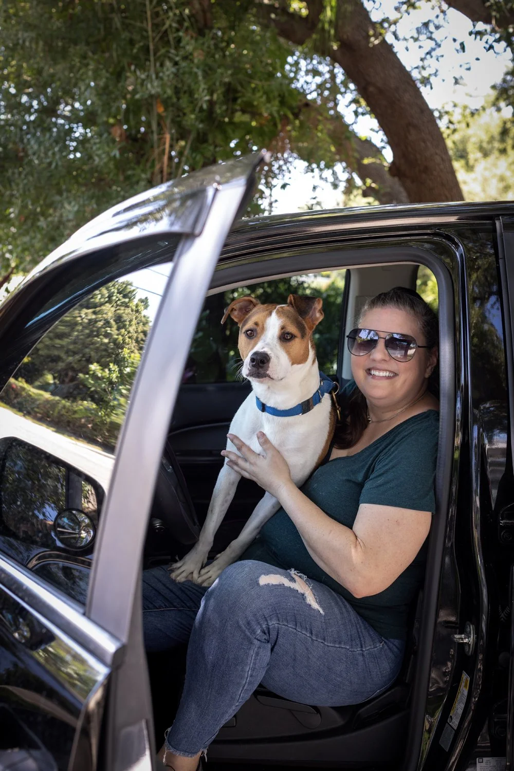 Woman with sunglasses smiling, sitting in car's passenger seat, holding a dog with a blue collar. The car door is open, and trees and greenery are visible outside.