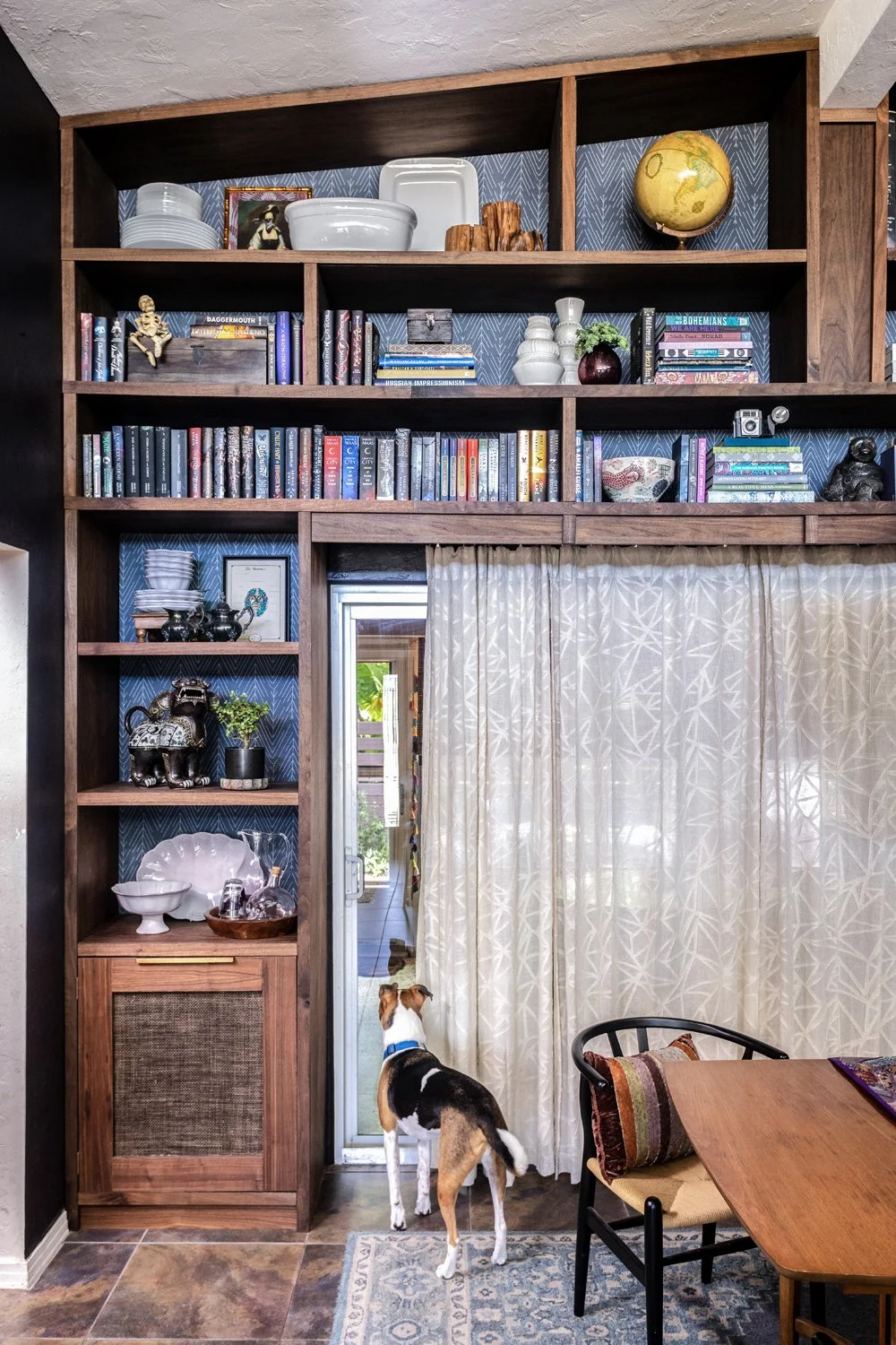 This image shows a cozy interior with a wooden bookshelf filled with books, decorative items, and ceramics. A small dog, possibly a Beagle mix, stands on a rug looking towards a sliding glass door covered with cream-colored patterned curtains. To the right of the dog, there is a table with a colorful cushion on a black chair, and the floor has a tile pattern.