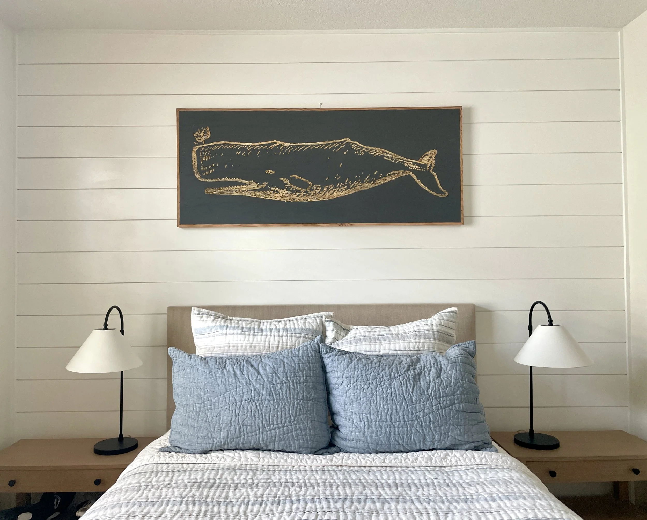 A bed with blue crinkled bedding and a white quilt, flanked by two black bedside lamps on wooden nightstands, against a white shiplap wall with framed whale artwork.