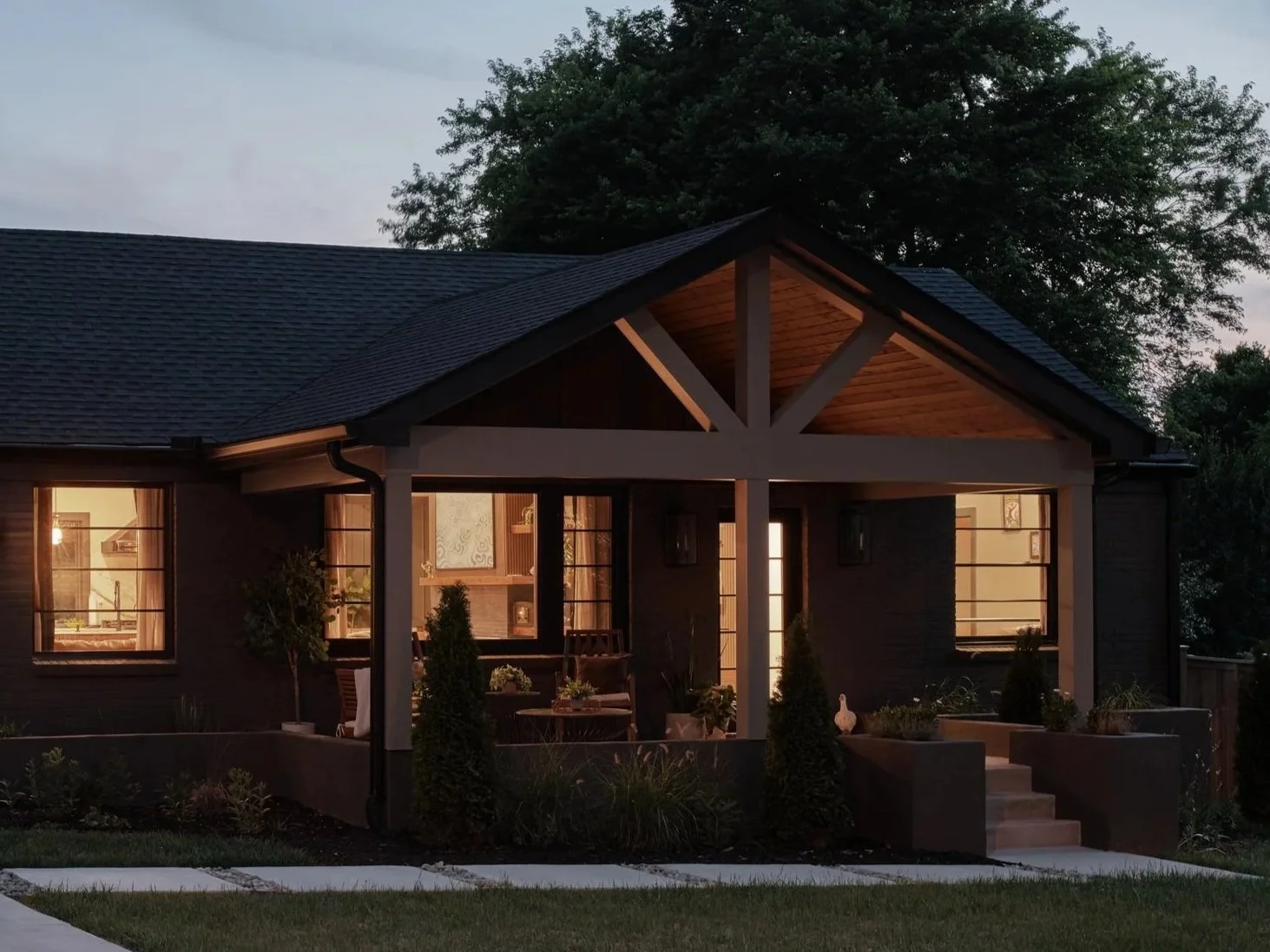 A suburban house at dusk with warm interior lighting, dark siding, a gabled roof with wooden accents, and a front porch decorated with plants and outdoor furniture.