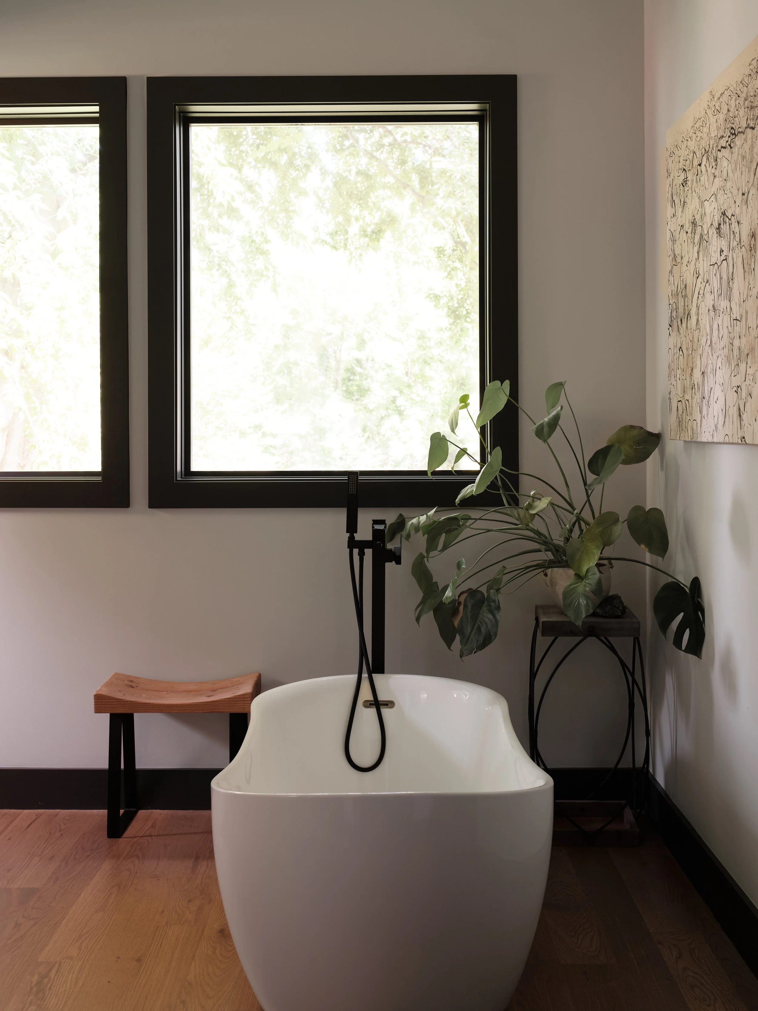Modern bathroom with a freestanding bathtub, two black-framed windows showing green trees outside, a wooden stool, a white chair, indoor plants, and a piece of abstract wall art.