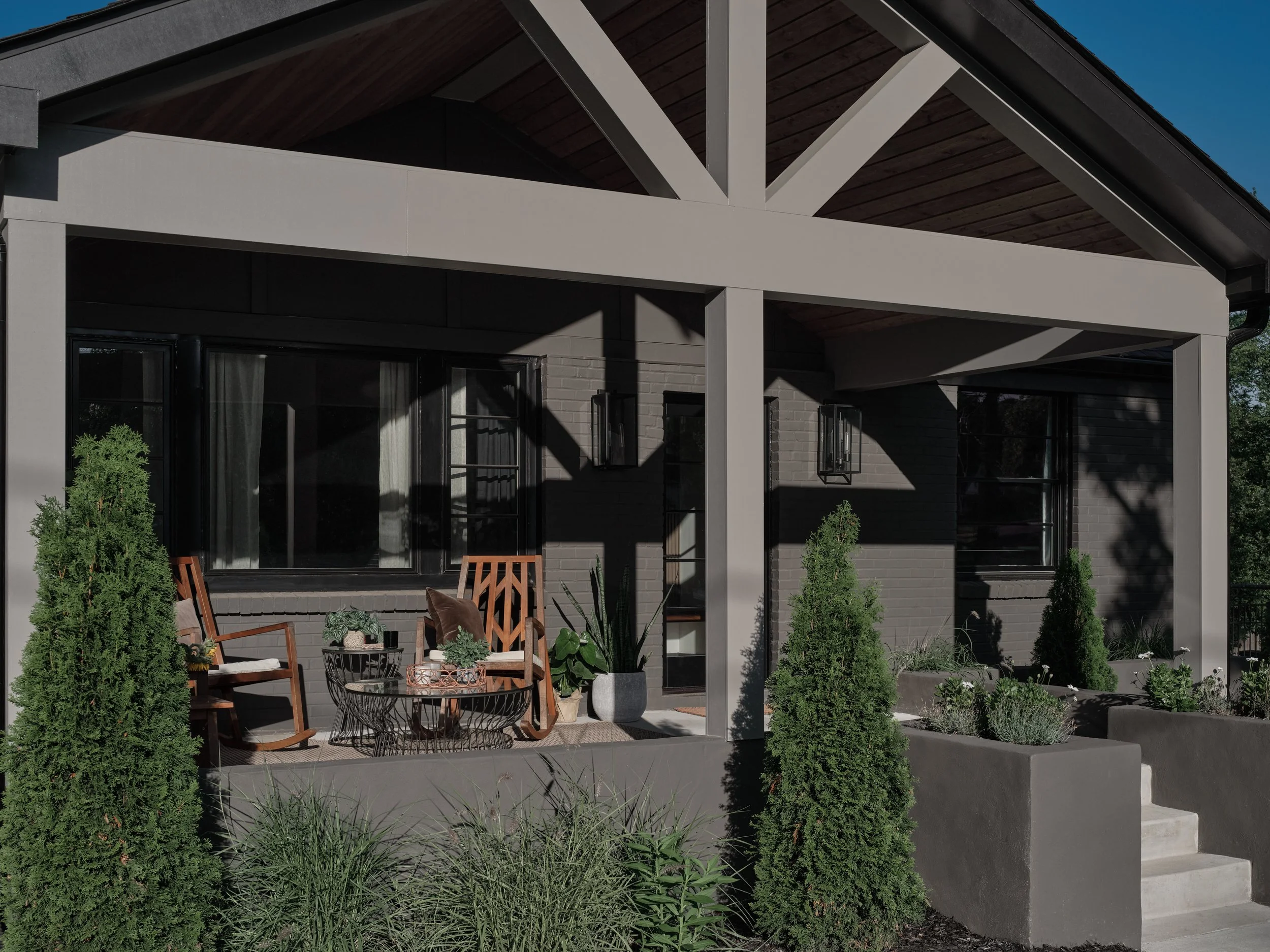 A modern house exterior with a patio, black-framed glass doors, wooden ceiling, and surrounding green plants.