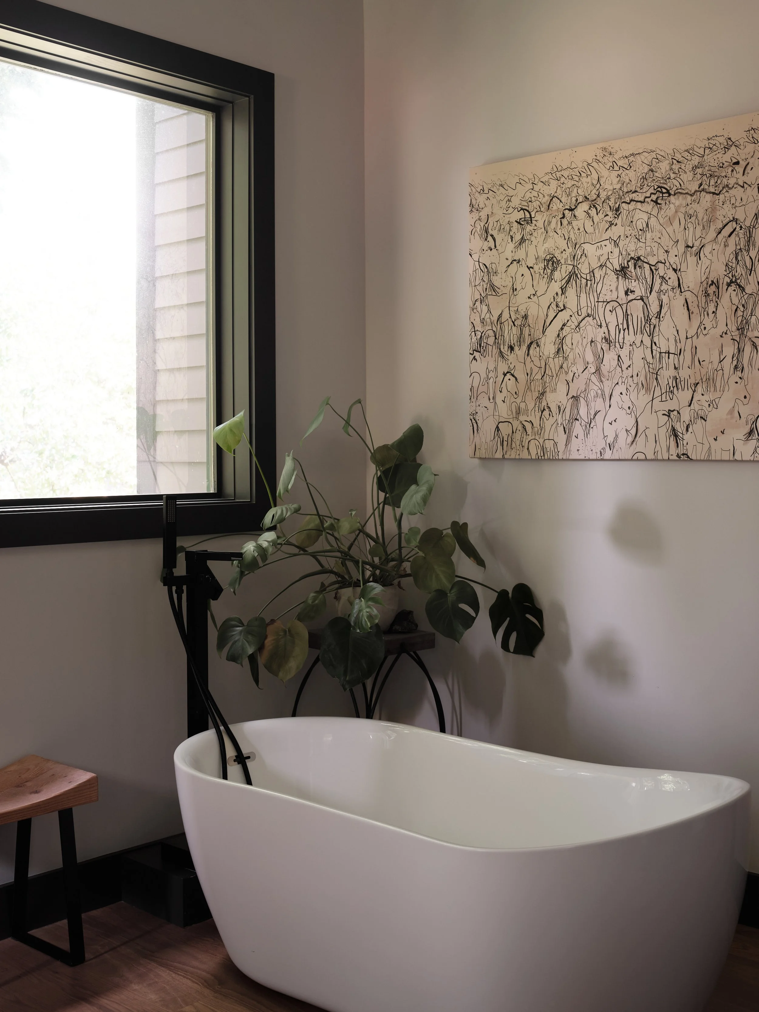 A modern bathroom with a white freestanding bathtub near a window, a wall-mounted art piece with black abstract line drawings, and a green houseplant on a small table.