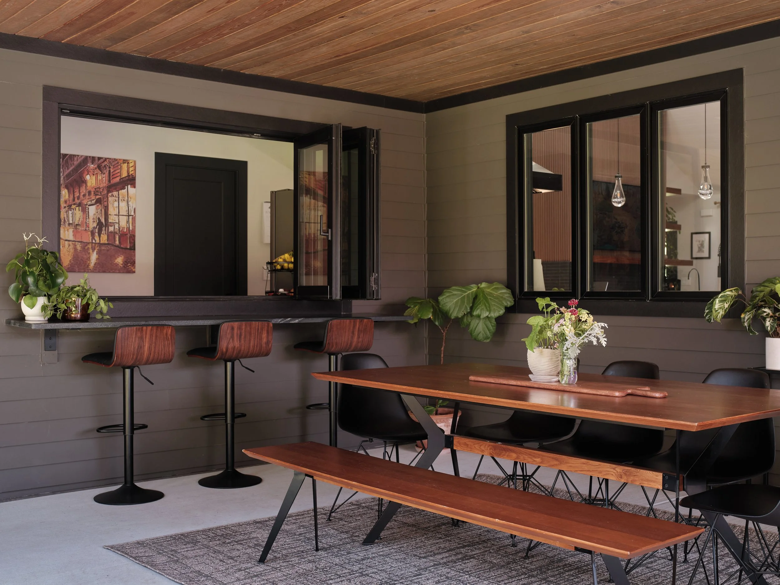 Modern dining area with a wooden table, black chairs, potted plants, wall art, and large windows with views of an interior space