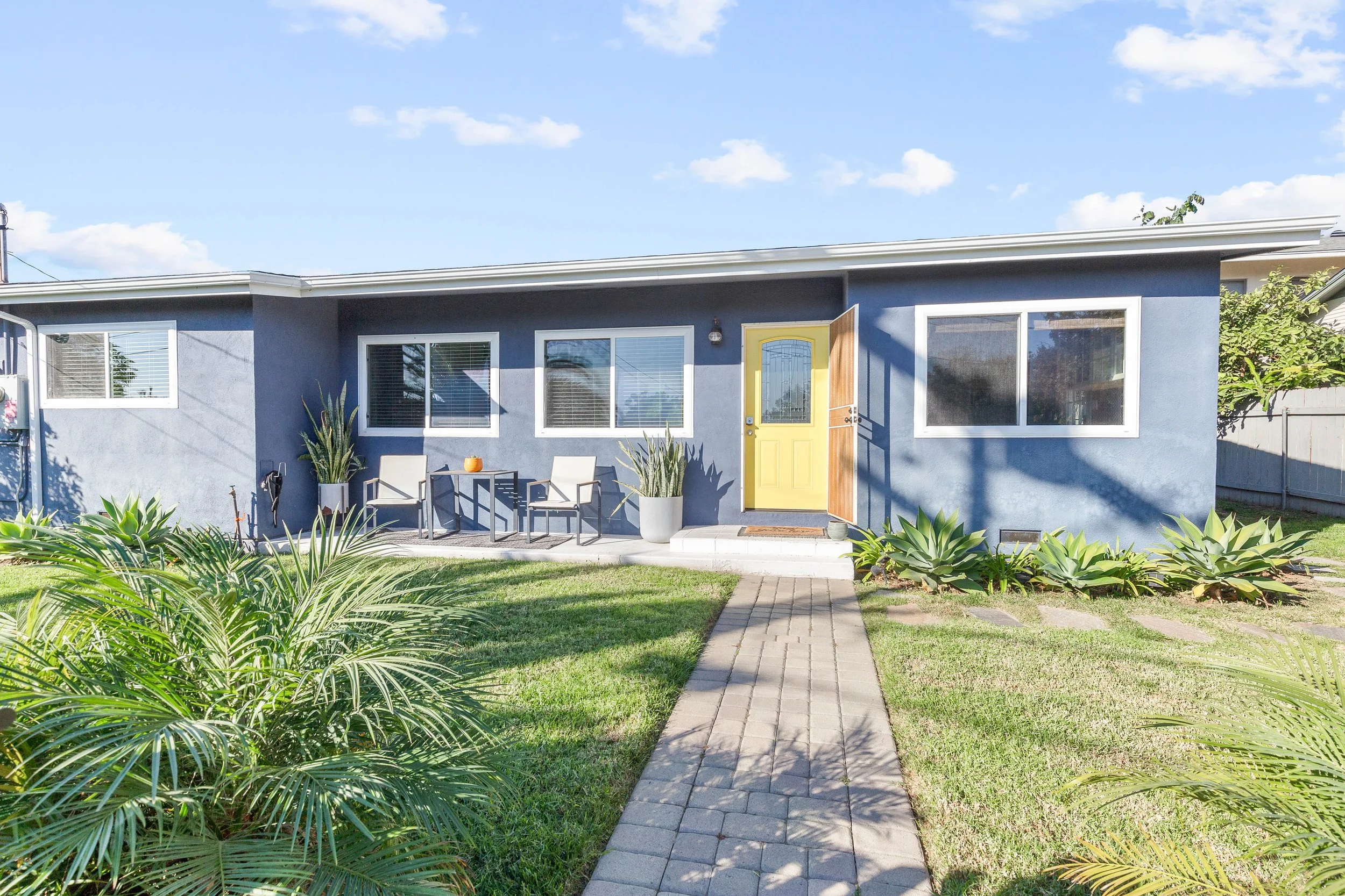 Front view of a blue house with a yellow front door, two large windows, a small sidewalk, outdoor chairs, potted plants, and a well-maintained lawn with lush greenery under a partly cloudy sky.