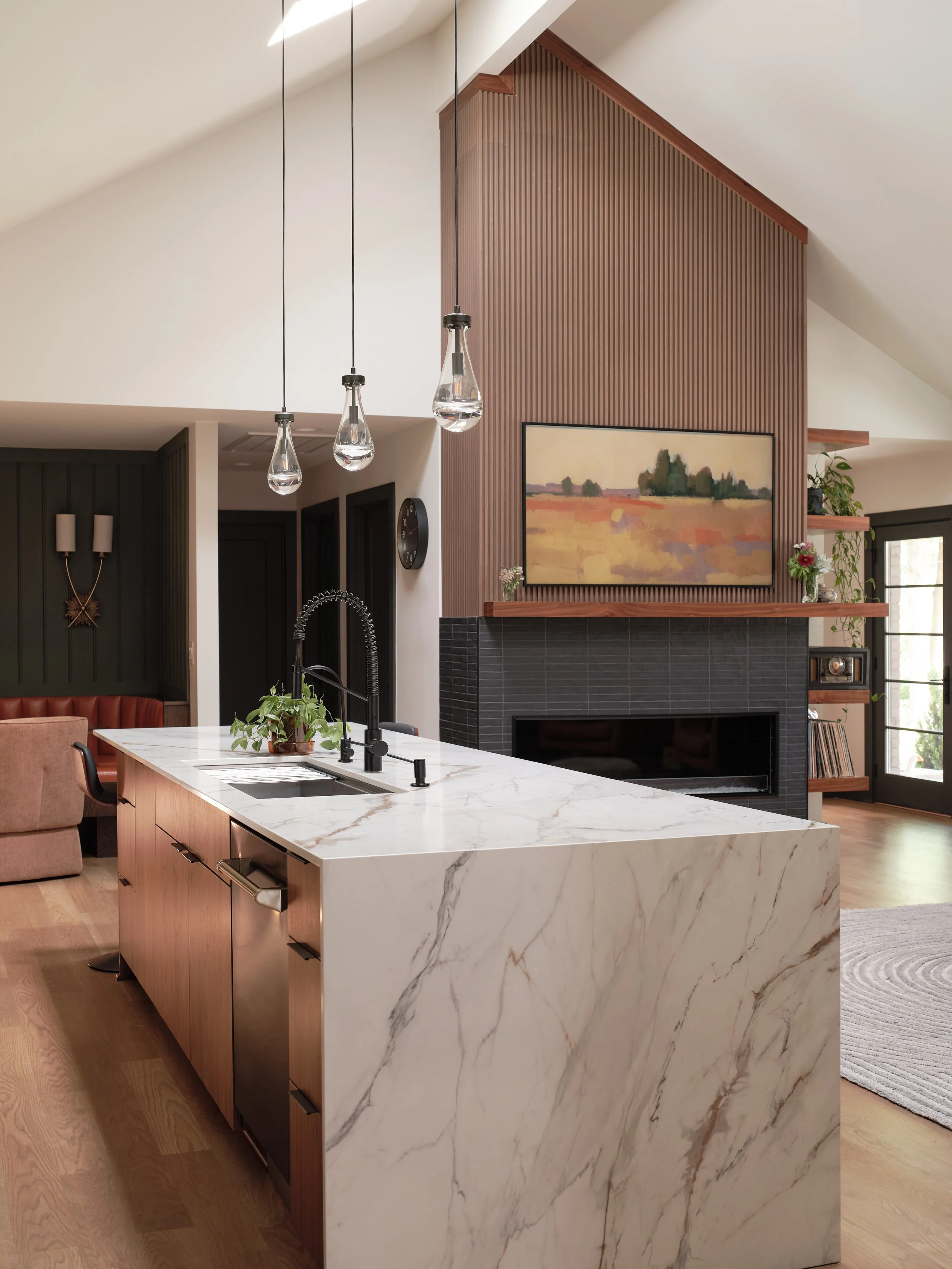 Modern kitchen with a white marble island, black sink, and a black faucet. Pendant lights hanging above the island, wooden cabinetry, and a painting above the fireplace. A plant and small decorative items on the mantel, with a tapestry of a field sce
