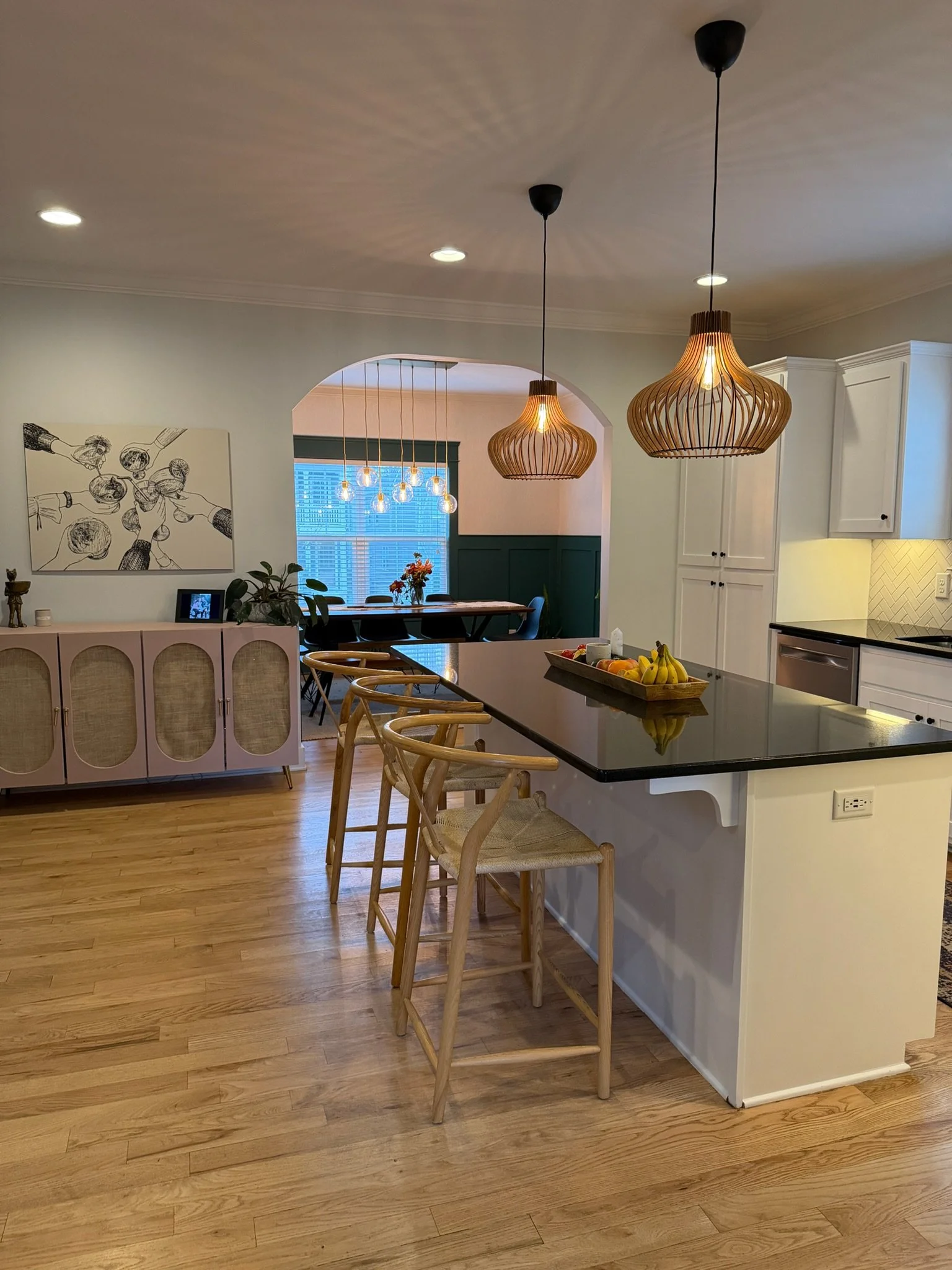 The image shows a kitchen and dining area with two wooden pendant lights hanging from the ceiling, a black countertop, white cabinetry, and a fruit bowl on the counter. The dining area includes a table with chairs, and a window with blinds in the bac