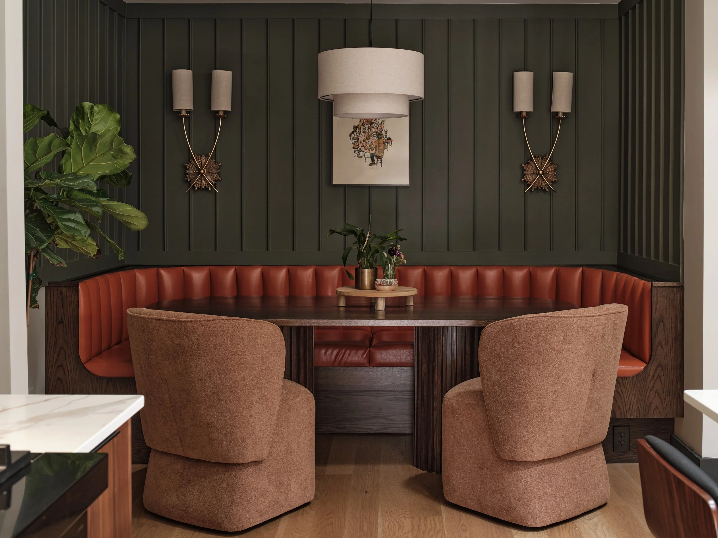 A cozy dining nook with a dark green paneled wall, a semi-circular red leather banquette, a wooden table, and upholstered chairs. Decor includes a small potted plant on the table, a large wall art piece, and coordinated wall sconces with beige shades