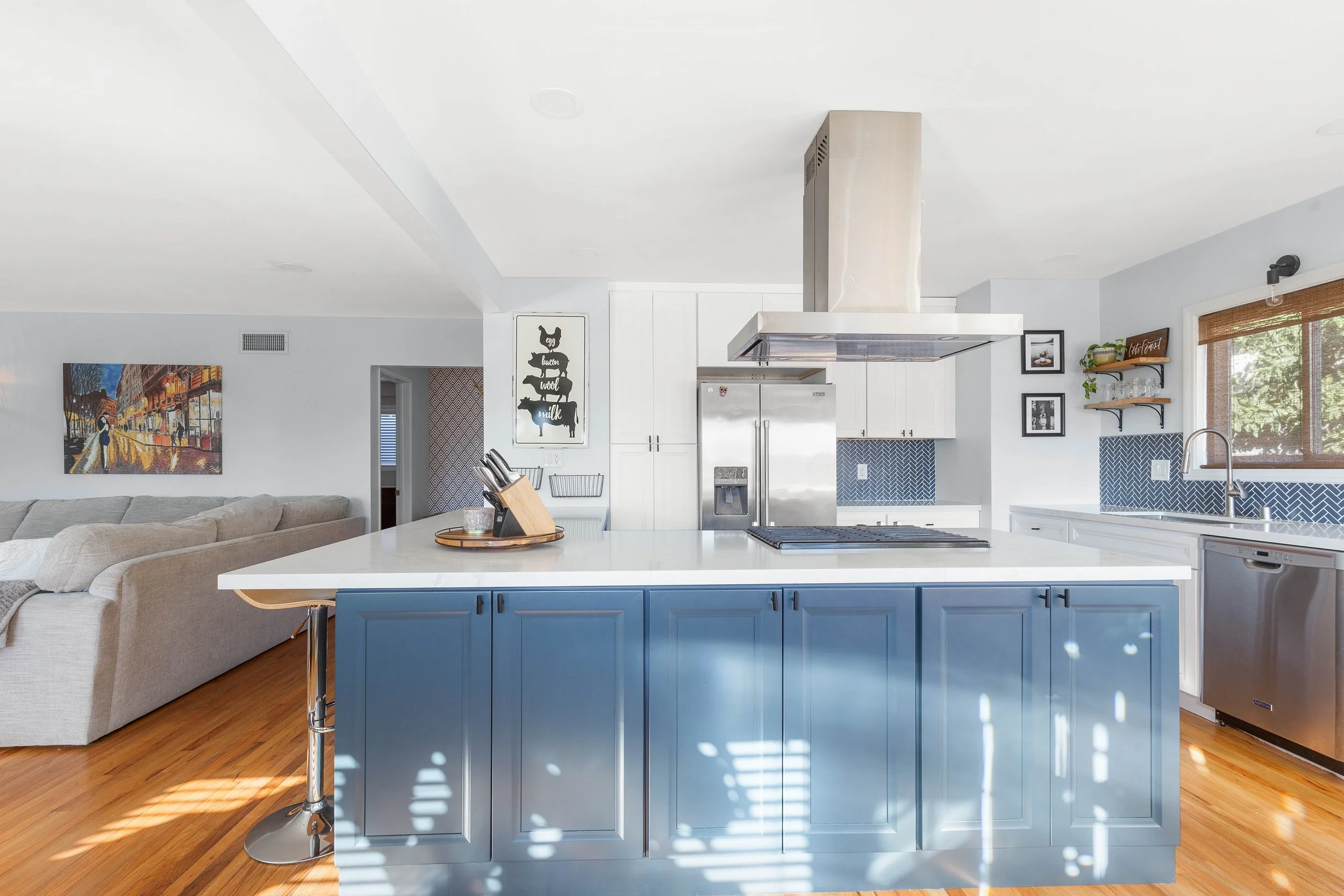 Modern open-concept kitchen with a blue island, stainless steel range hood, white cabinets, and a living area with artwork on the wall.