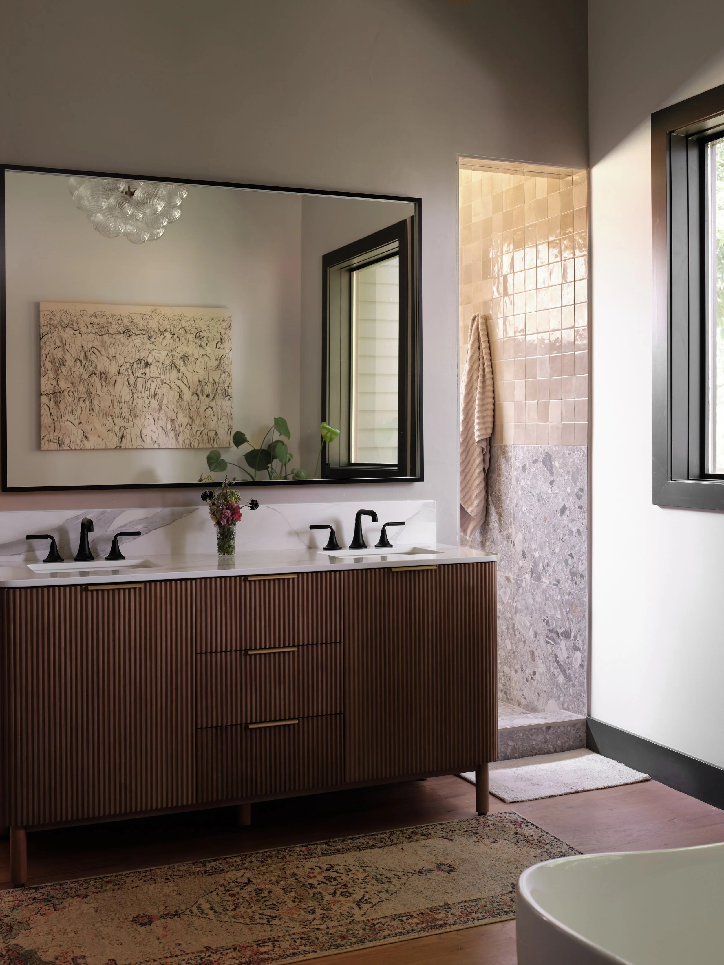 A modern bathroom with a wooden vanity, black fixtures, and a large mirror. There is a window with black framing and a tiled shower area with a towel hanging on the wall. A bathtub is partially visible in the foreground, with a beige rug on the woode