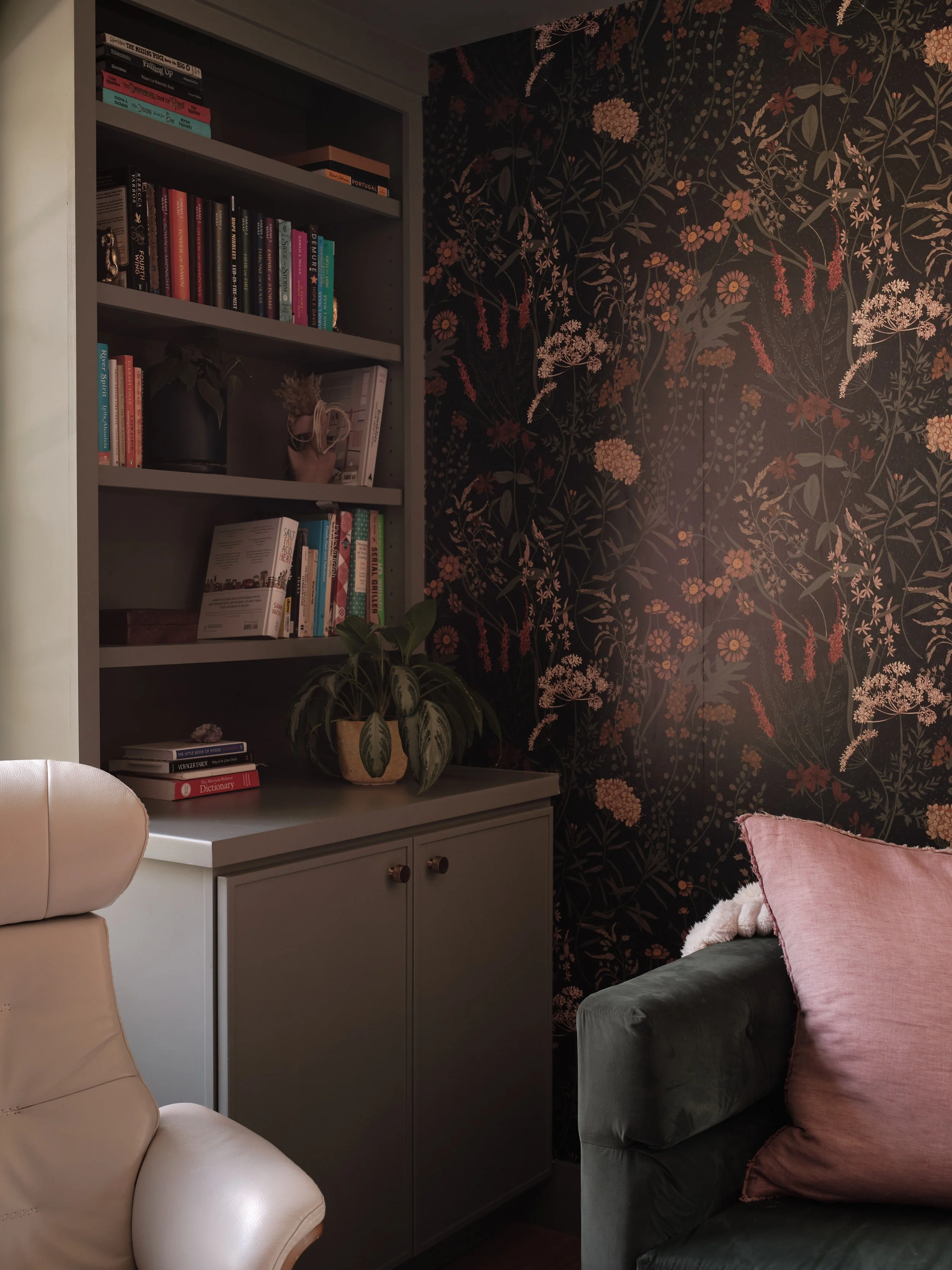 A corner of a room featuring a bookshelf filled with books, a small plant on top of a light-colored cabinet, dark floral wallpaper, and part of a gray sofa with a pink pillow.