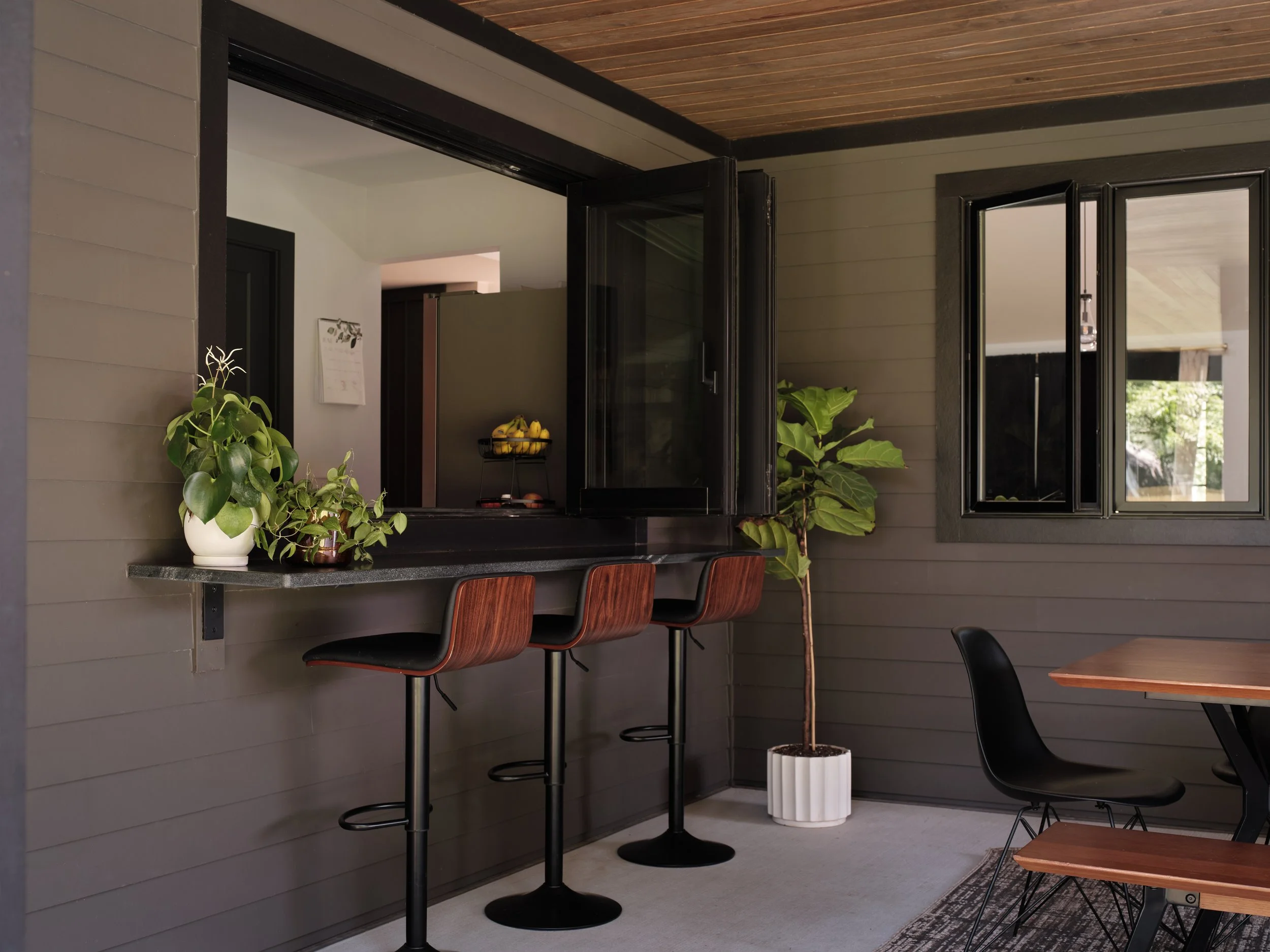 Interior of a modern cafe or kitchen area with a gray wall, a black counter, and a large window. There are green plants on the counter and a potted tree near the window. Bar stools are lined up along the counter and part of a wooden table with a blac