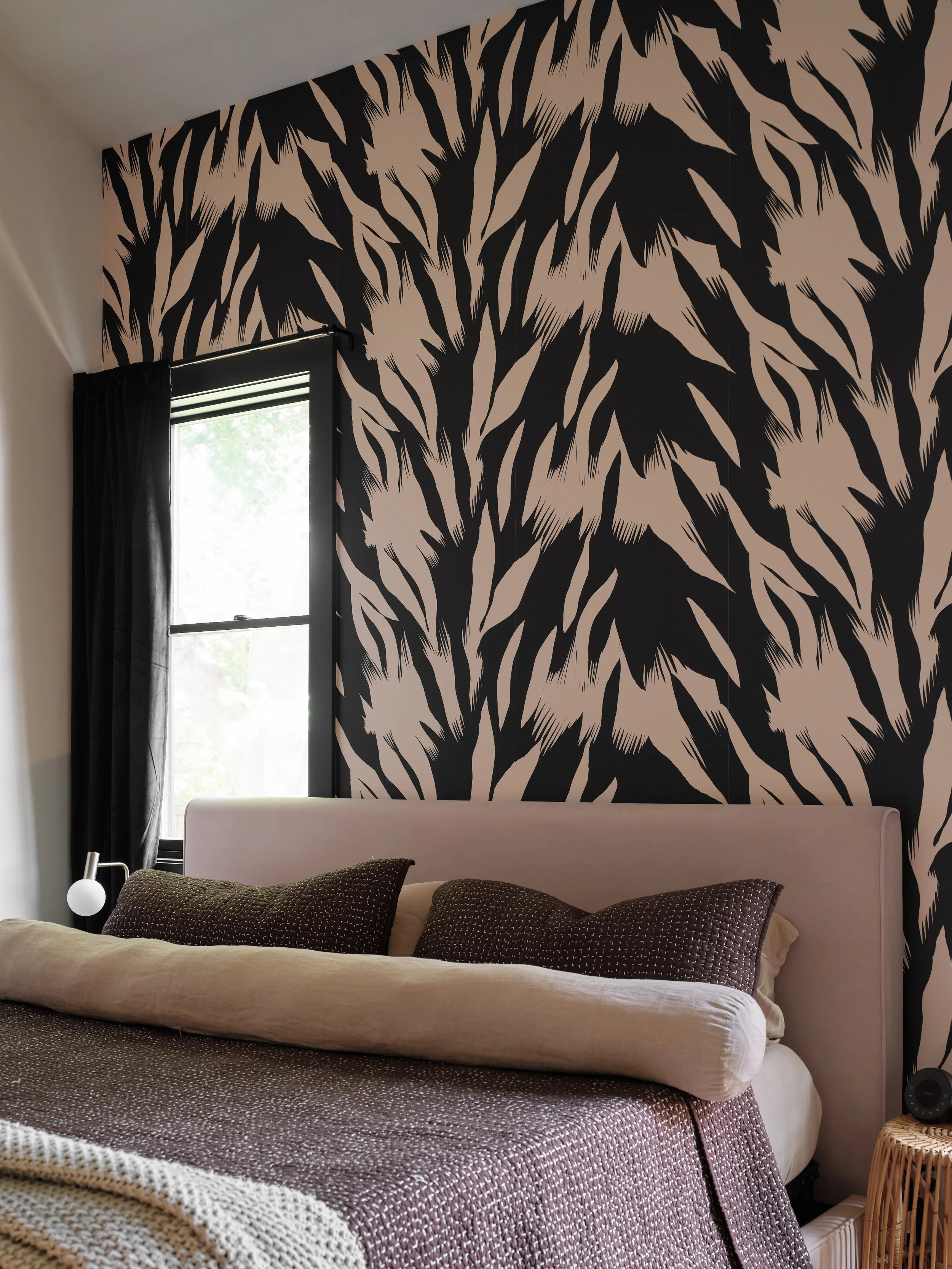 A bedroom with a large black and beige abstract leaf pattern wallpaper behind the bed. There is a window with black curtains, and the bed has a beige duvet with a textured brown blanket and dark brown pillows. A wicker side table with a clock and a s