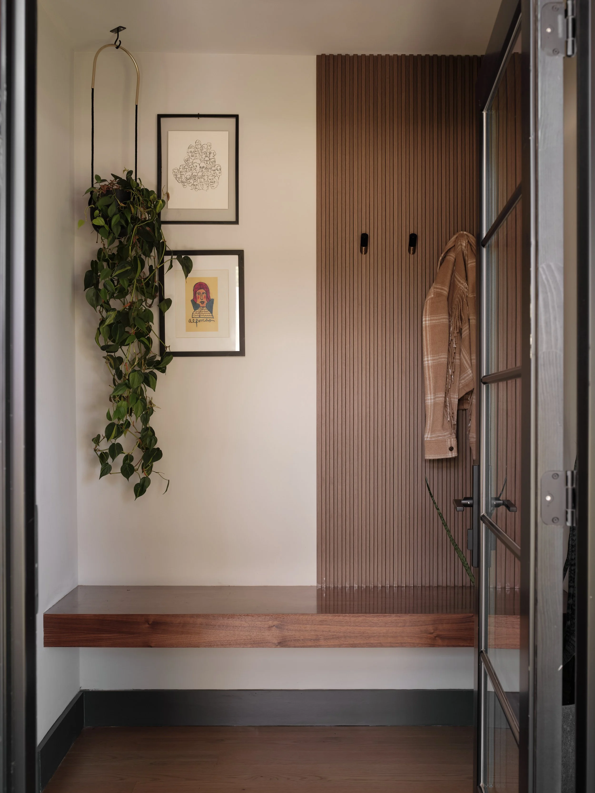 Inside an elevator with a wooden panel on one wall, two framed art pieces, a hanging potted plant, and a brown plaid coat hanging on the wall.