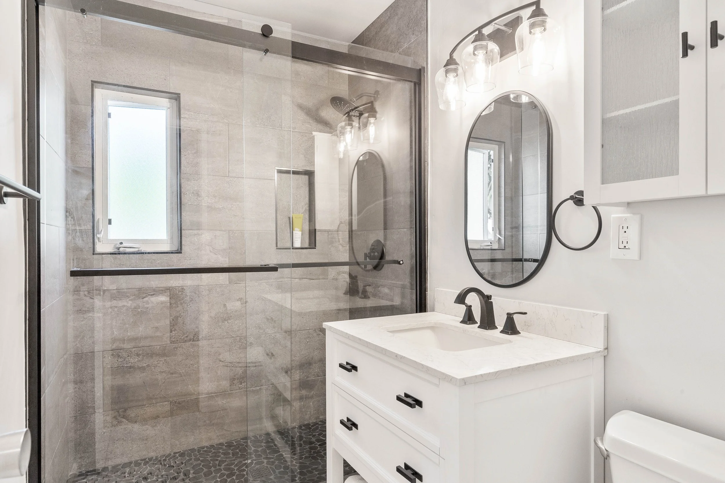 A modern bathroom with a glass-enclosed shower, a white vanity with a marble countertop, oval mirrors, and a black faucet and fixtures.