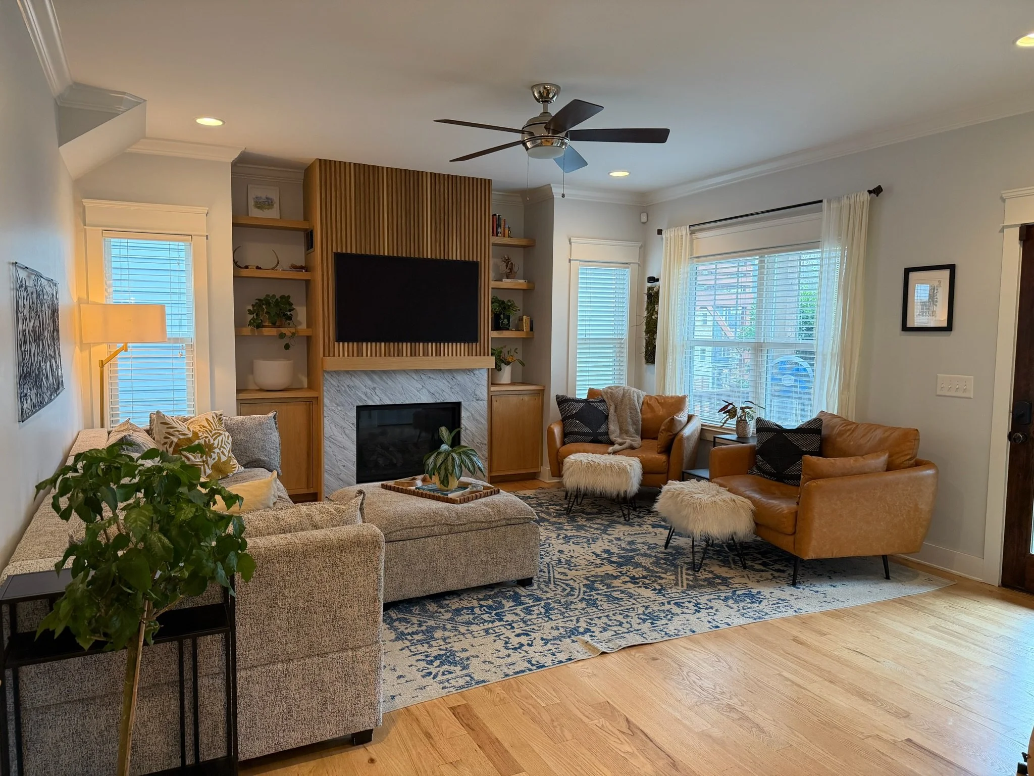 A living room with beige sofas, a patterned area rug, a fireplace with built-in shelves, a large window with curtains, plants, and a ceiling fan.