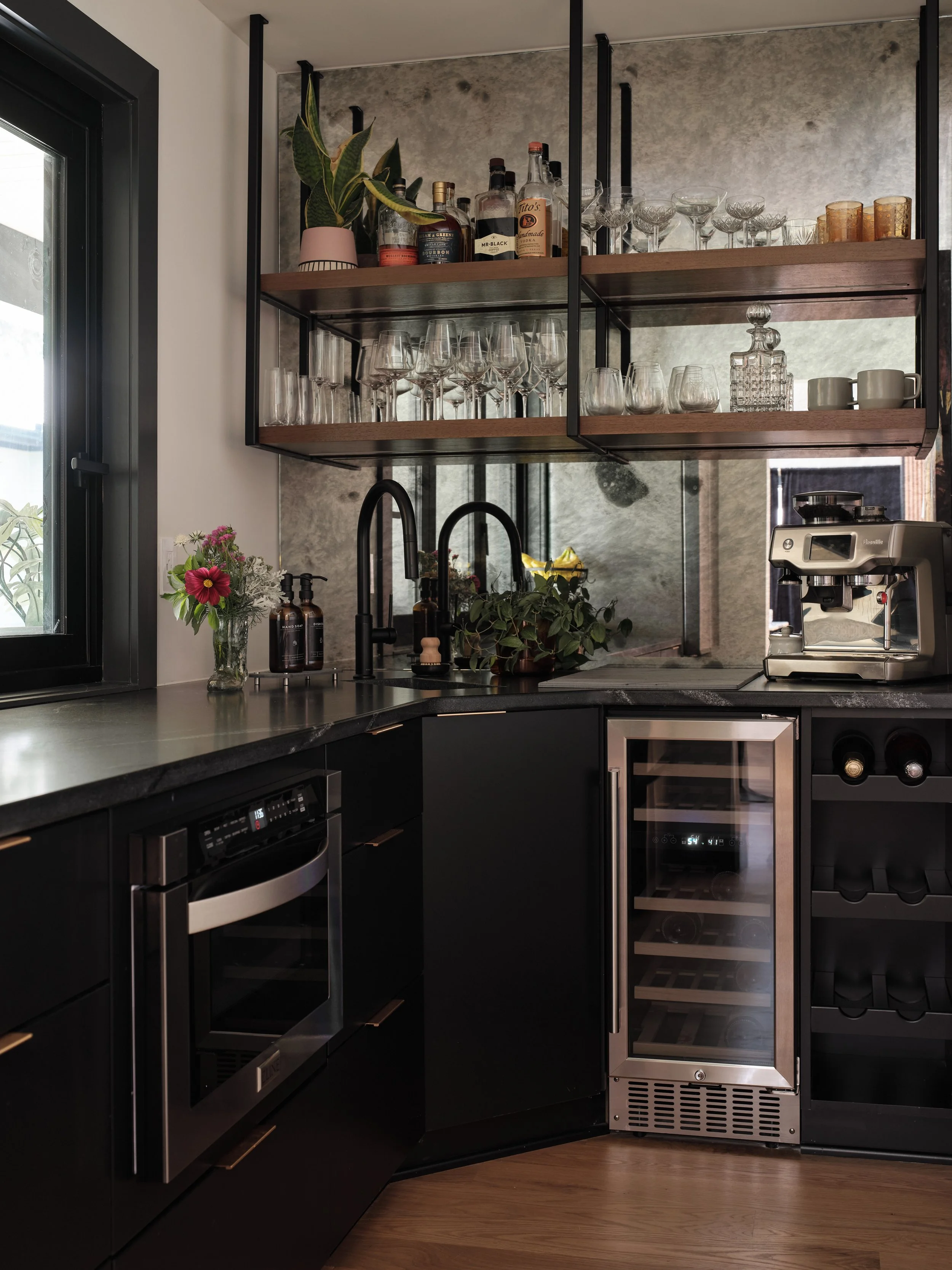 A modern kitchen with black cabinets, a black countertop, a wine cooler, and a kitchen shelf with glasses, bottles, and plants. A window with black trim is on the left, and there are various plants and kitchen appliances visible.