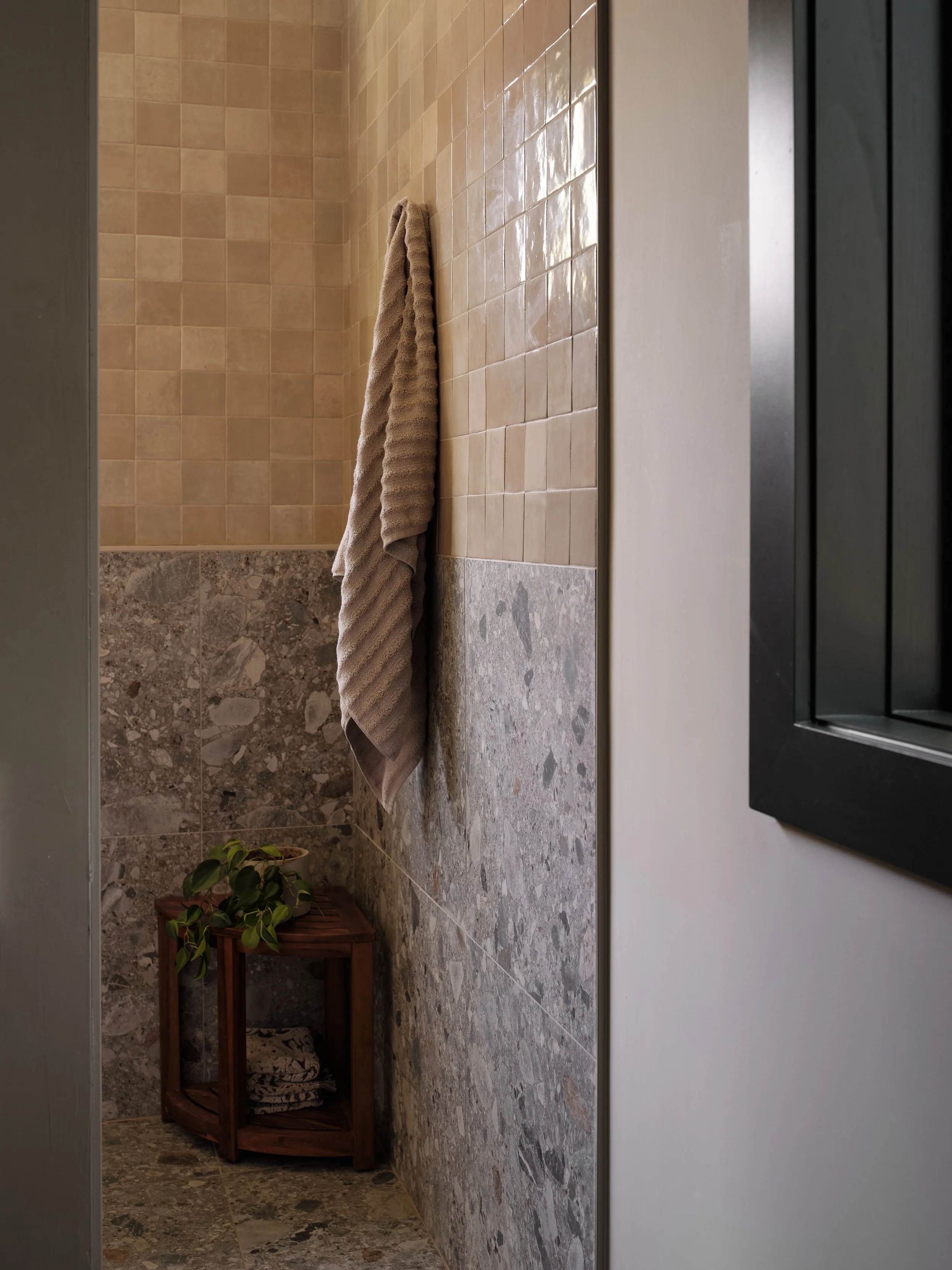 A small brown wooden shelf with a potted green plant and folded towels, against a wall with beige tiles and a marble floor, with part of a white wall and a dark doorframe visible.