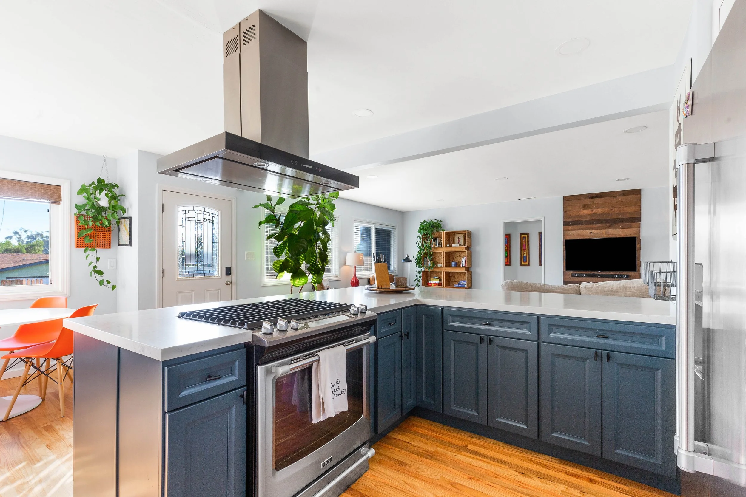 Bright kitchen with blue cabinets, white countertops, stainless steel oven and range hood, green plants, living room with TV and wooden shelves, windows with blinds.