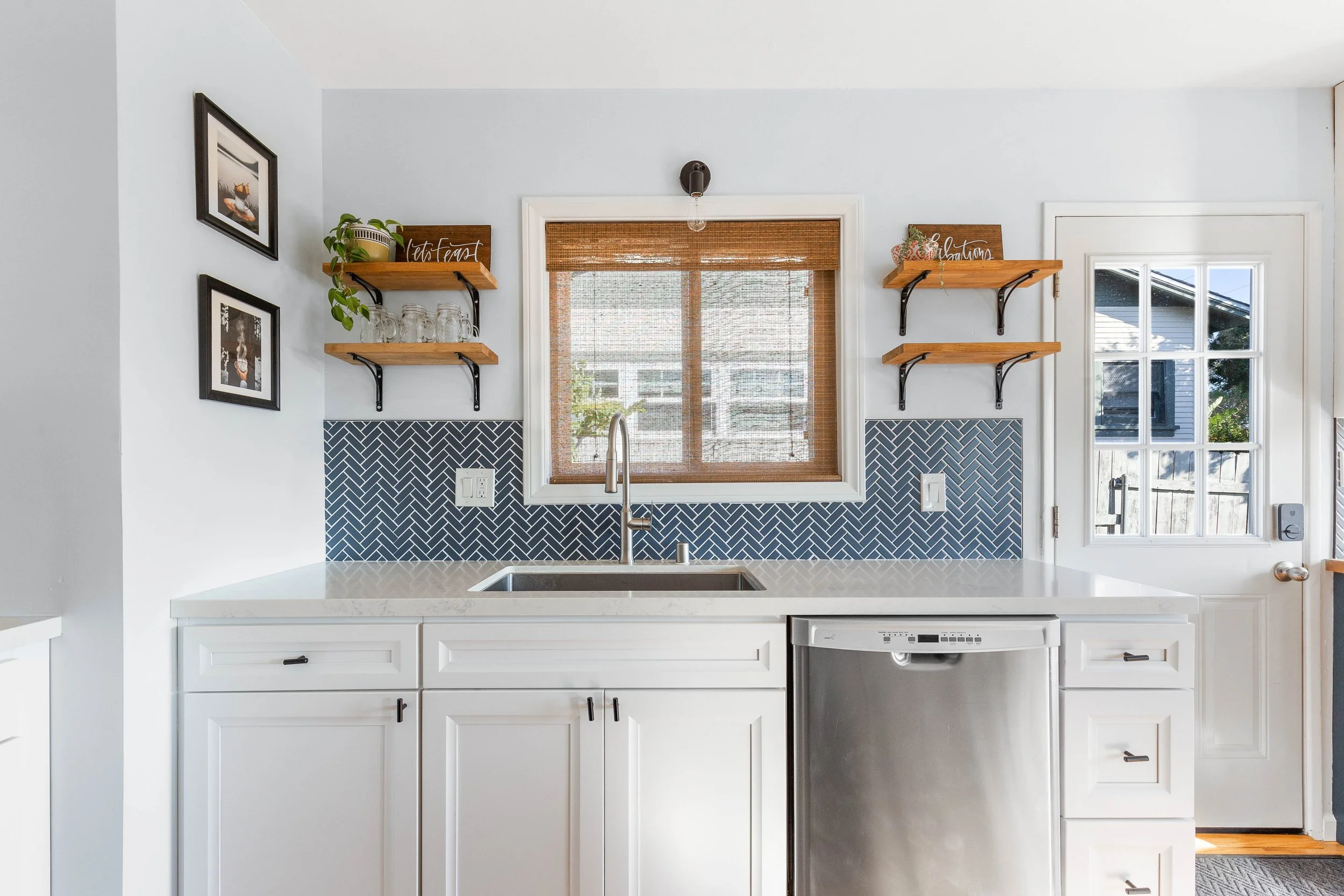 A kitchen with white cabinets, a white countertop, a stainless steel sink, a dishwasher, and a blue herringbone tile backsplash. There are two wooden shelves with black brackets, one with jars and a plant, and the other with decorative signs. A windo