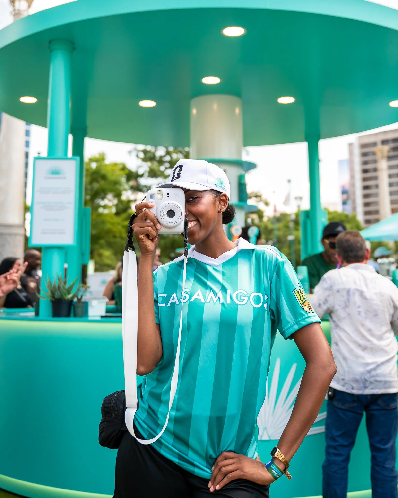 A woman wearing a white cap and turquoise sports jersey taking a photo with a Polaroid camera in an outdoor setting with a large teal and white structure in the background.