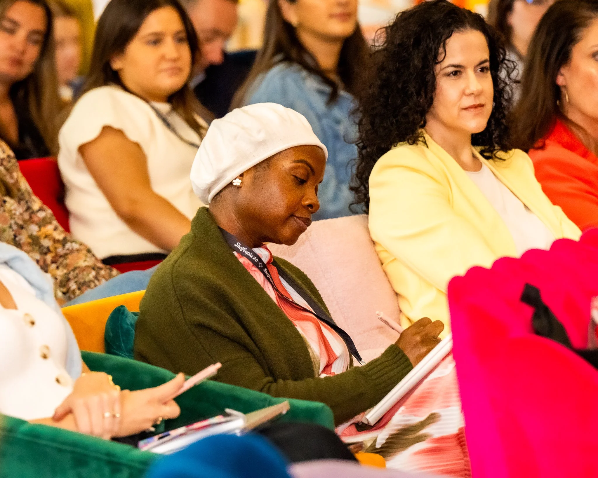 A group of women seated in an audience, some taking notes, attending an indoor event or conference.
