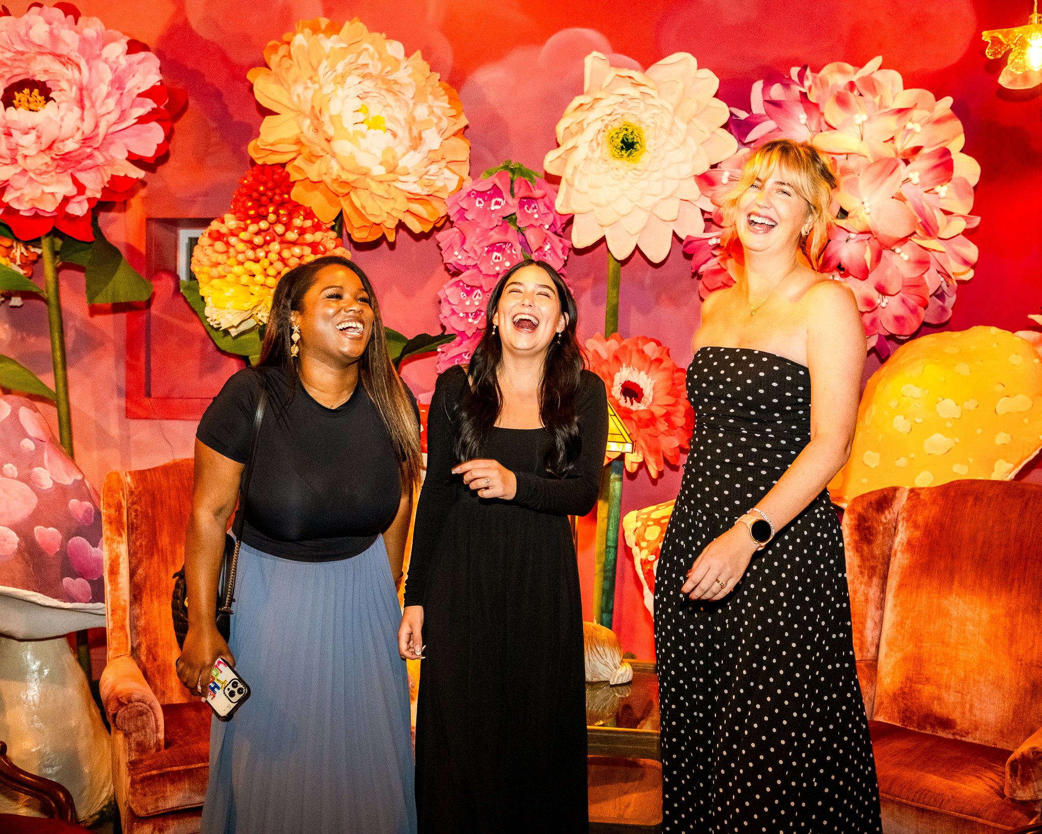 Three women are standing together and smiling in front of a colorful background with large flowers and pink walls.