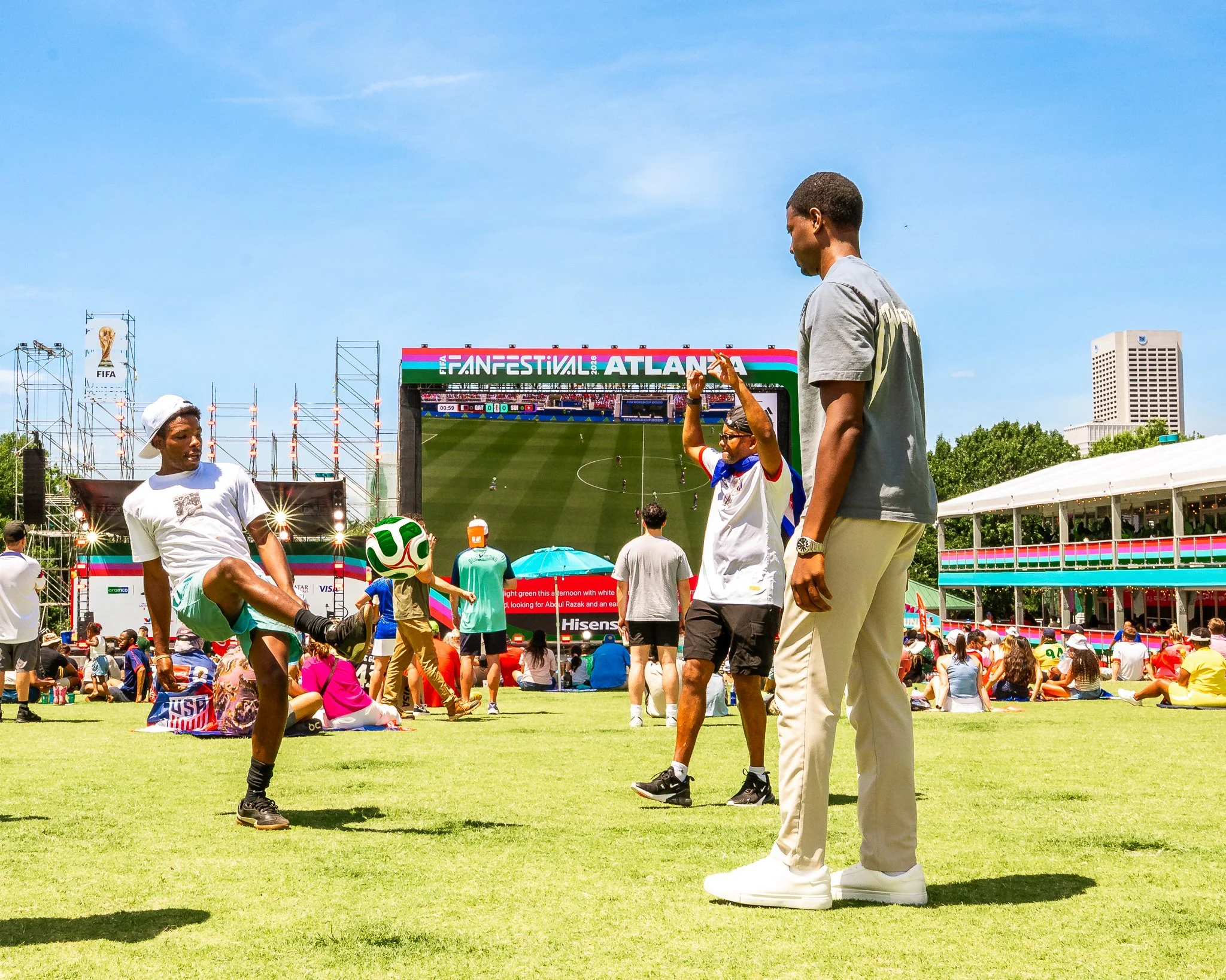 People watching a soccer match at an outdoor stadium with a large screen displaying the game, surrounded by a crowd under a clear blue sky.