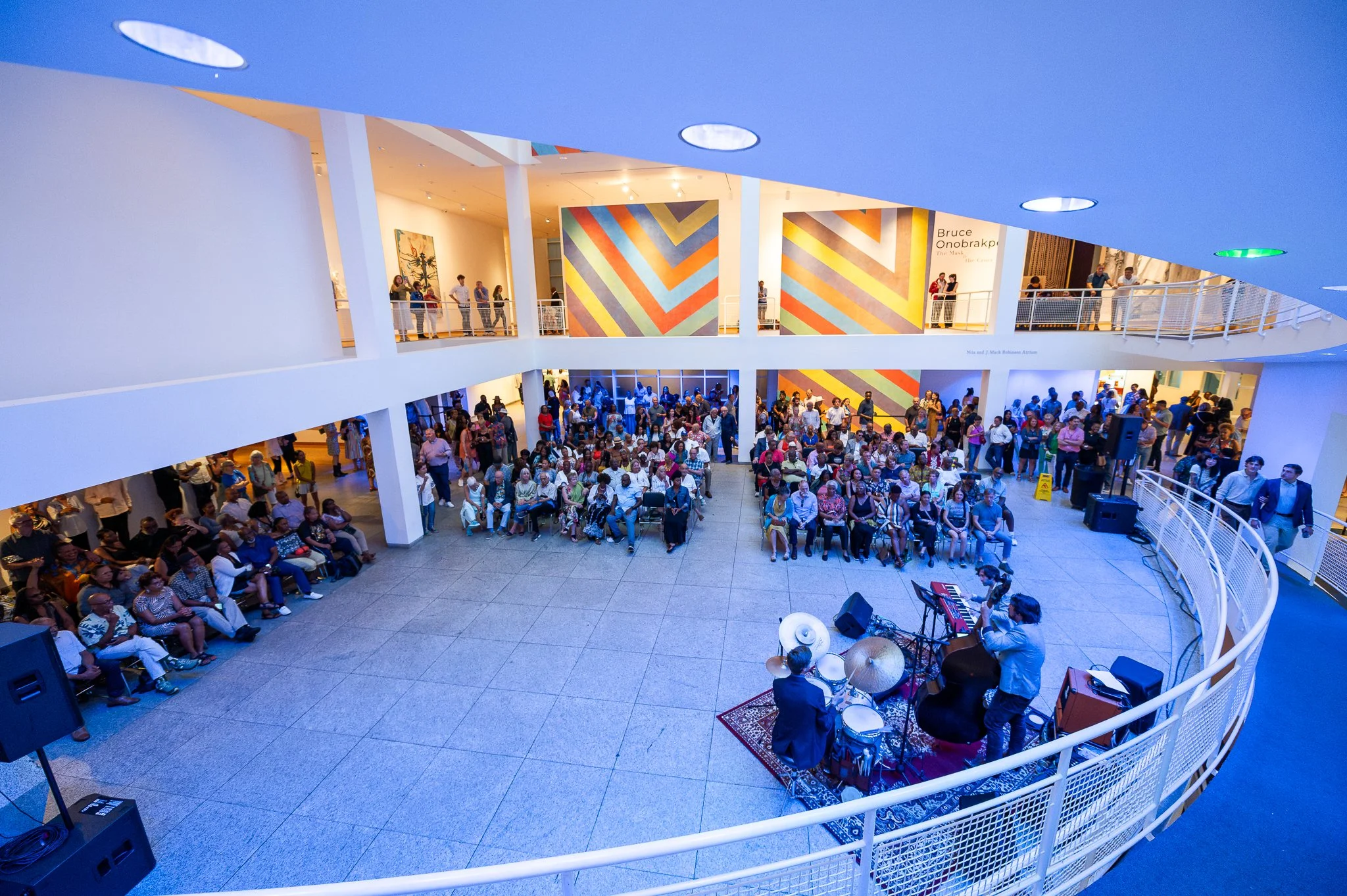 An indoor music performance with an audience seated in front of a band playing drums, keyboard, and other instruments. The venue has multiple levels with viewers looking down and up at the performance. Colorful artwork is displayed on the walls.
