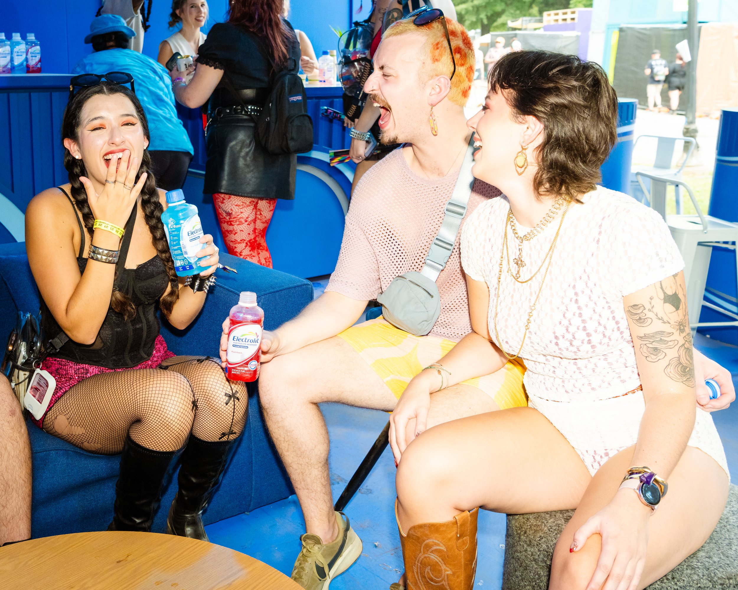 Group of young adults laughing and talking at an outdoor event with blue booths in the background. They are dressed in casual and colorful clothing, with one person holding Electrolit