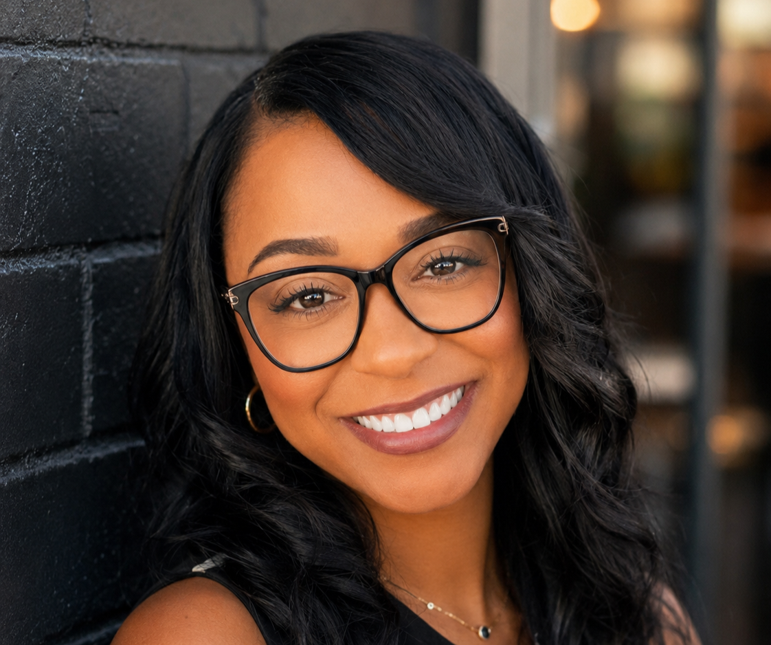 Close-up of a woman with black wavy hair, glasses, and earrings, smiling while leaning against a dark wall.
