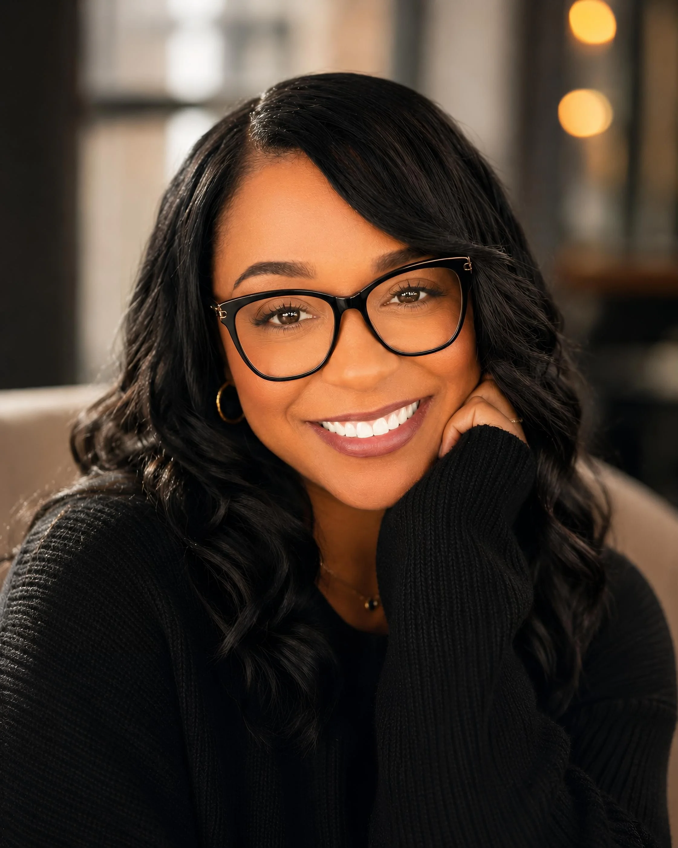 A woman with dark hair, glasses, and a bright smile, wearing a black sweater, in a warmly lit indoor setting.