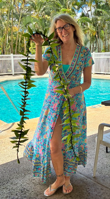 Woman in a colorful dress holding a long leafy branch by a swimming pool with a white fence and palm trees in the background.