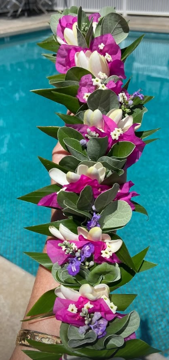 A hand holding a floral lei made of purple, white, and pink flowers with green leaves, against a swimming pool background.