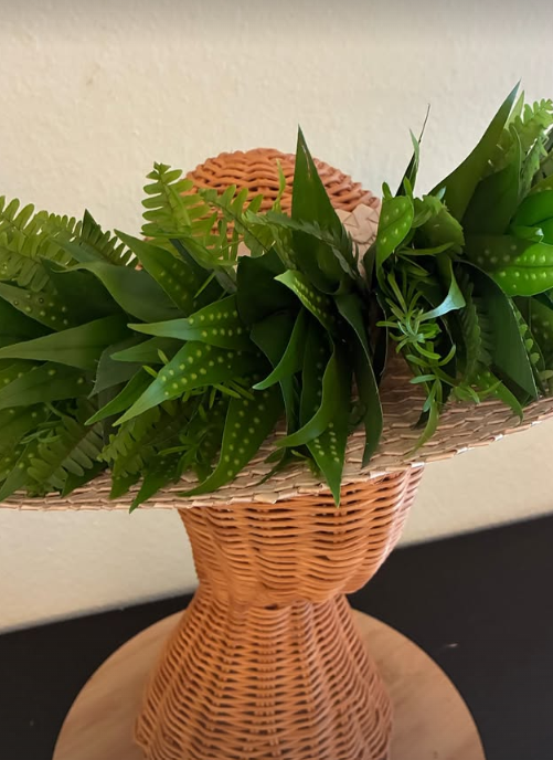 A woven basket filled with various green plants and leaves, placed on a table against a plain wall.