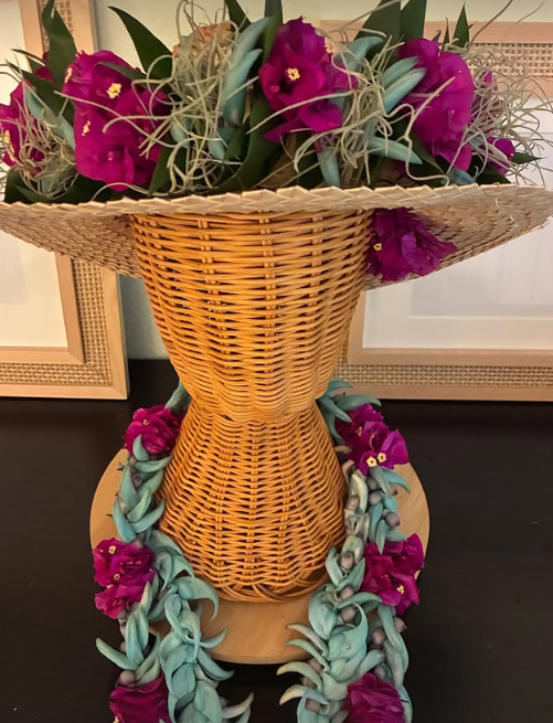 Wicker vase filled with purple flowers and greenery, with braided blue and green lei draped around it and on the surface beneath, set on a wooden table against a background of white cabinets.