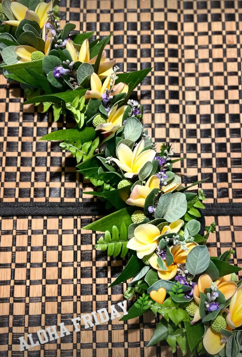 A floral lei made of yellow and purple flowers with green leaves, resting on a bamboo mat background with a "Aloha Friday" message