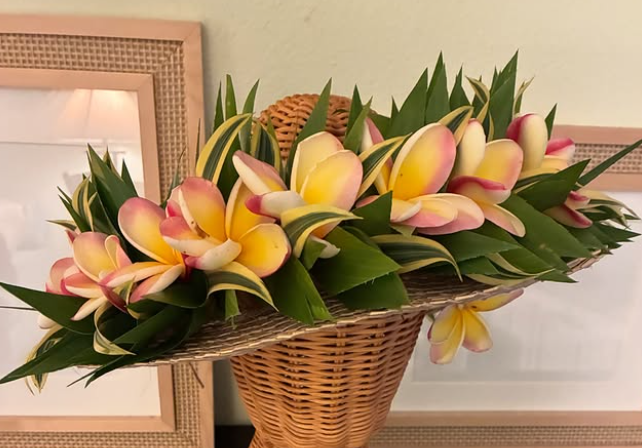 Wicker basket filled with yellow and pink plumeria flowers and green leaves, placed on a surface with picture frames in the background.